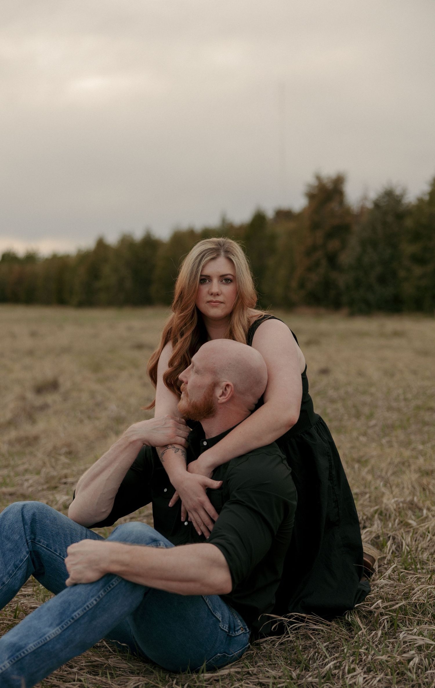 Couple sitting in a grassy field at dusk, woman embracing man from behind intimately.