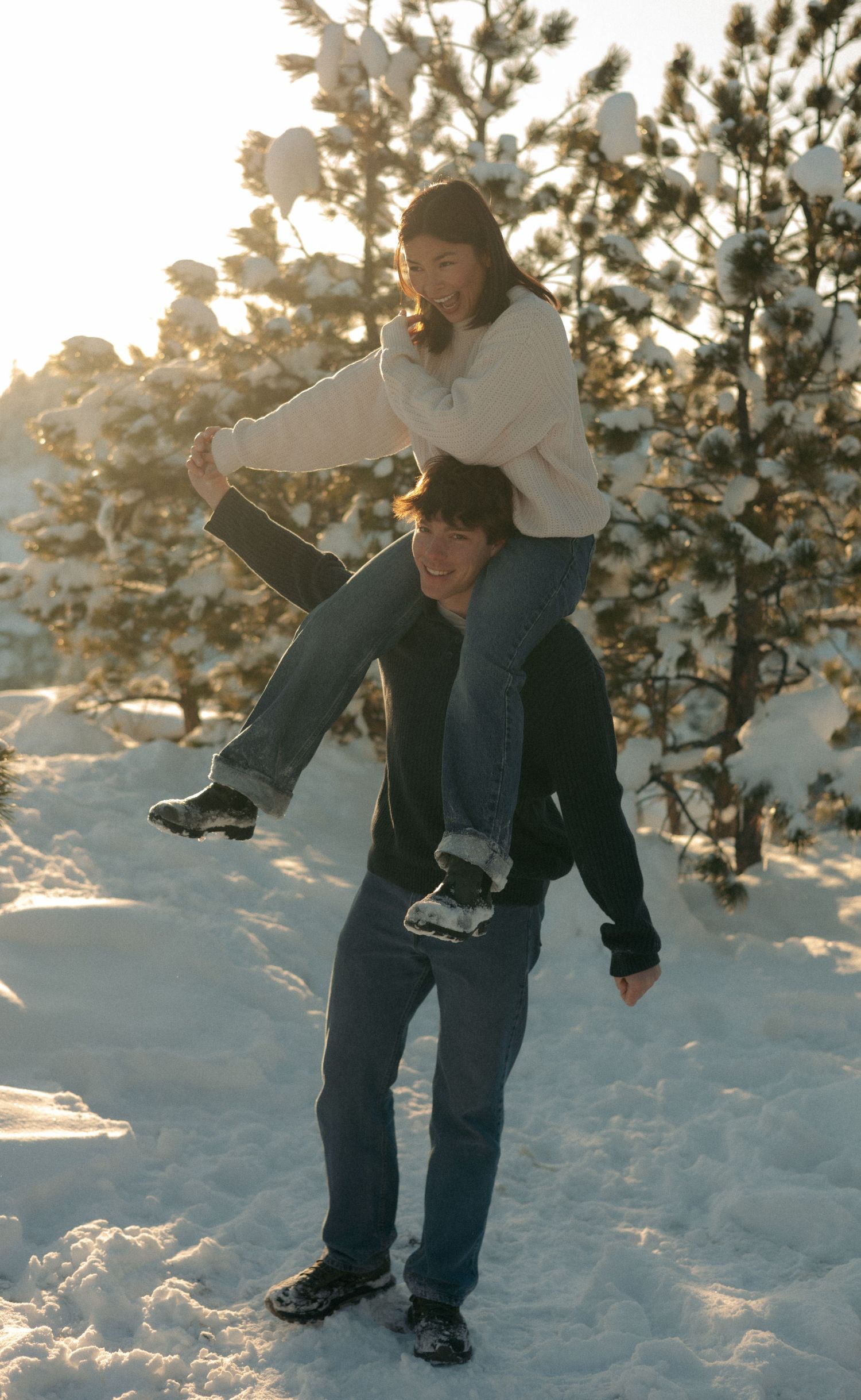 Couple playing in snow, woman on man's shoulders, surrounded by snow-covered pine trees in golden winter sunlight.