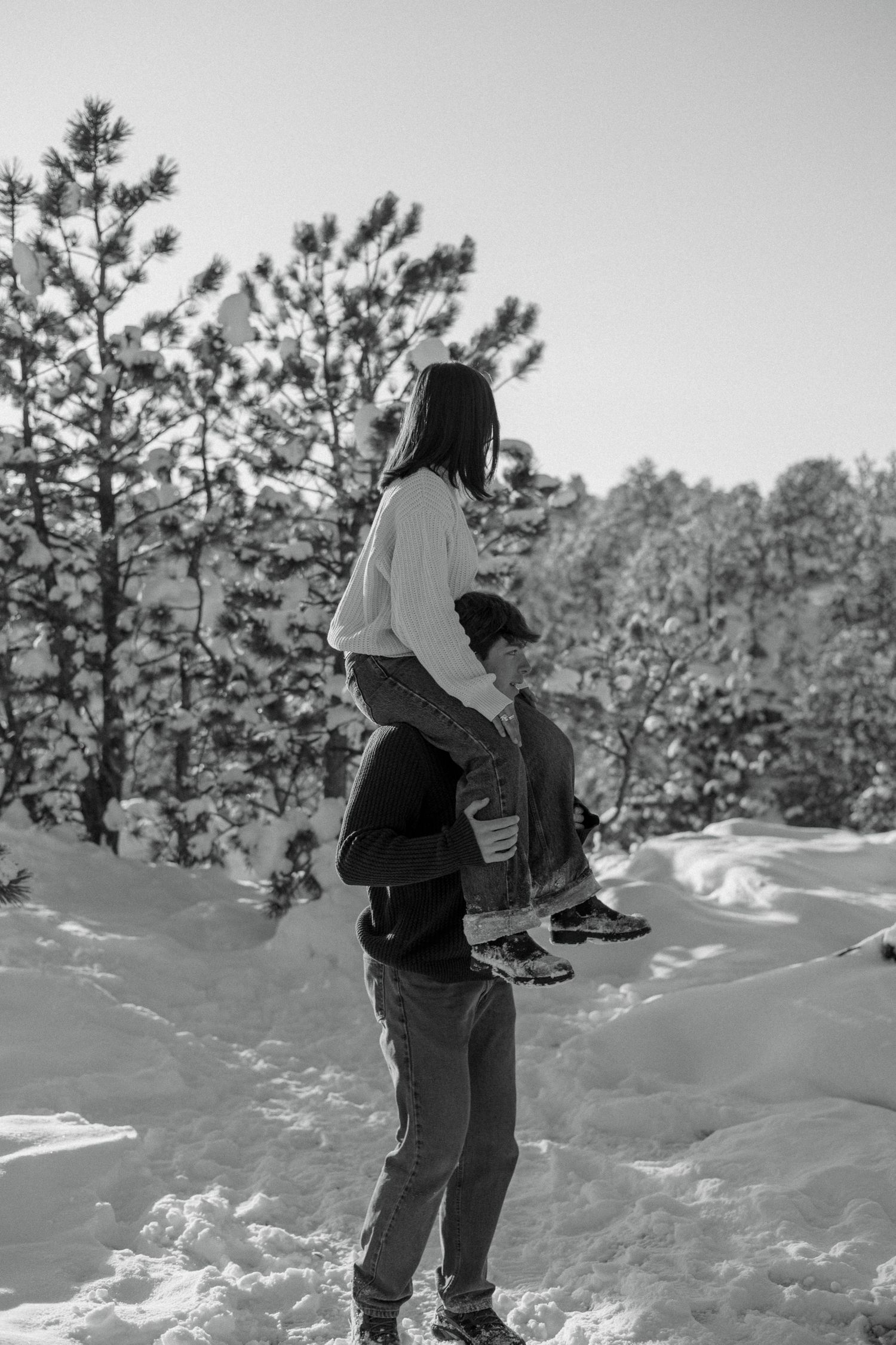Couple in snowy winter forest, woman sitting on mans shoulders surrounded by snow-covered pine trees.