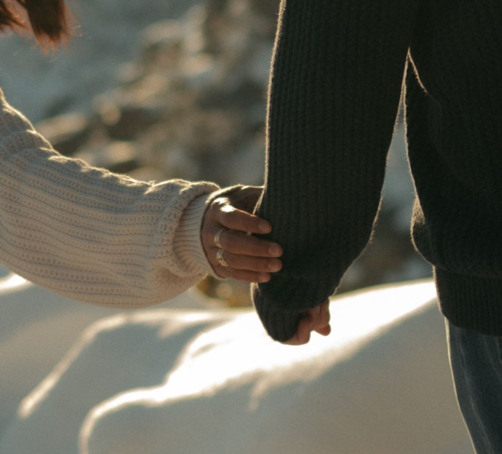 Close-up of two people holding hands while wearing cozy sweaters in bright winter sunlight outdoors.