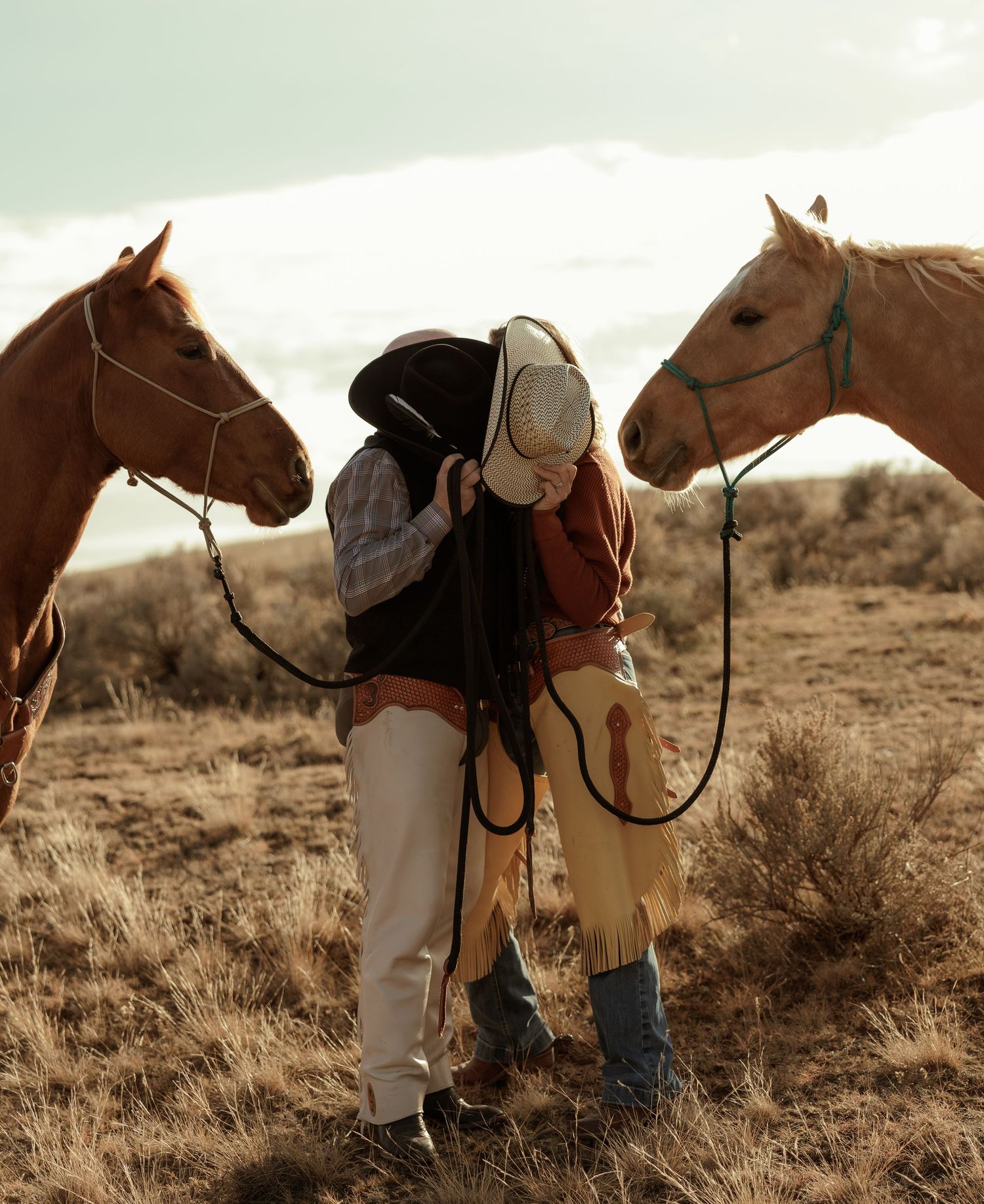 Couple sharing a kiss between two chestnut horses in a dry golden grassland landscape.