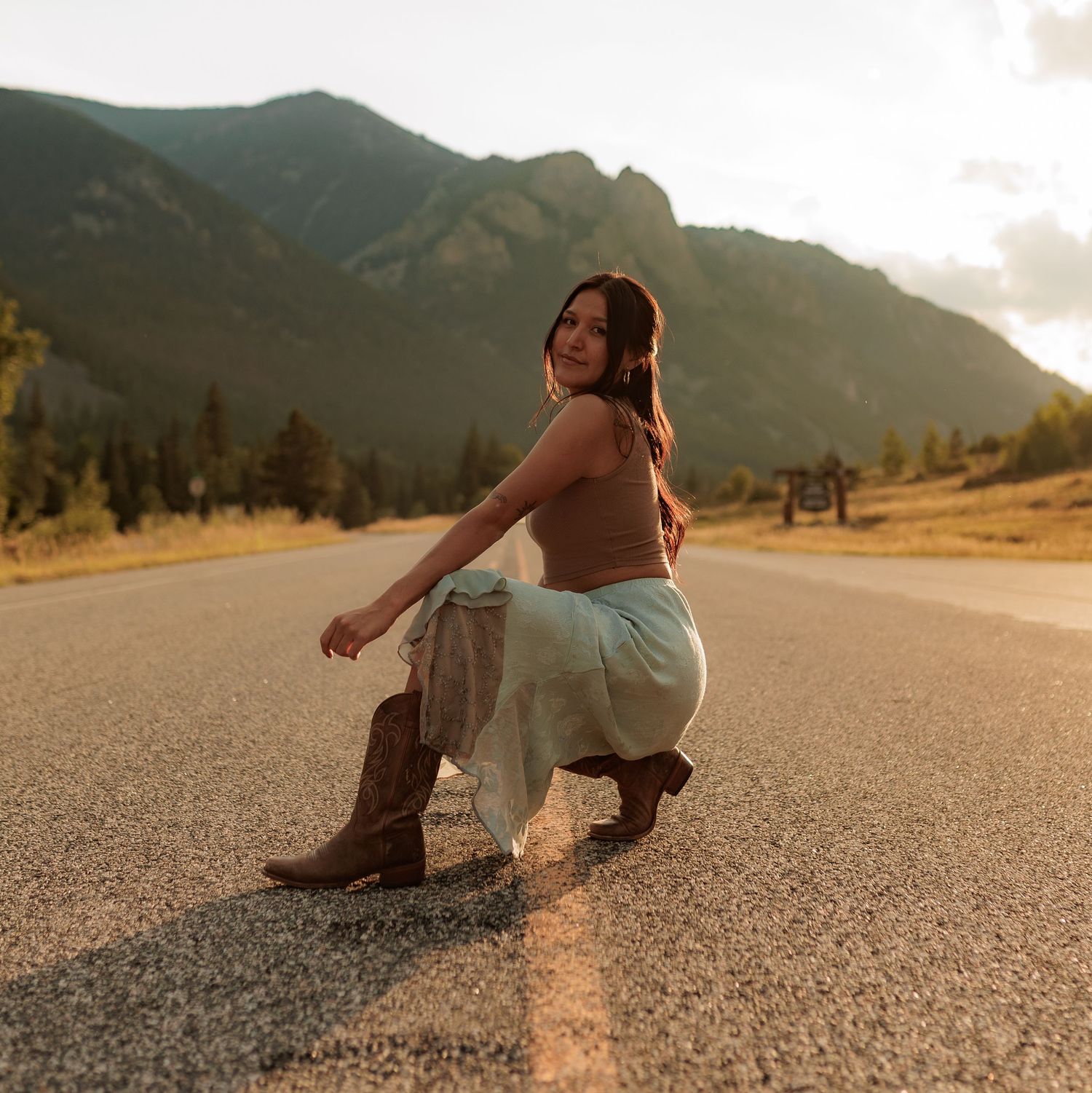 Woman in cowboy boots and skirt posing on a mountain road at golden hour in Colorado.