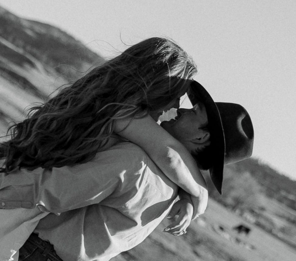 Black and white photo of a couple embracing and kissing outdoors, woman with long hair, man wearing a cowboy hat.