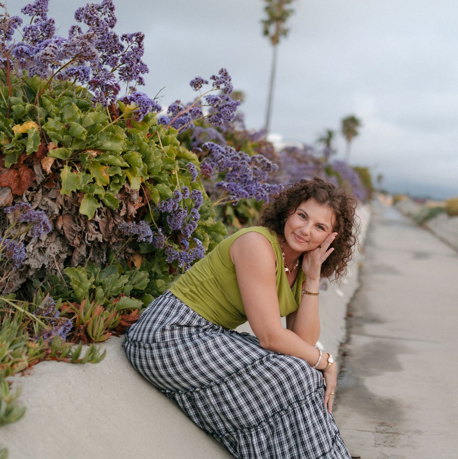 Woman in green top and plaid skirt sitting by purple wildflowers on a coastal pathway.
