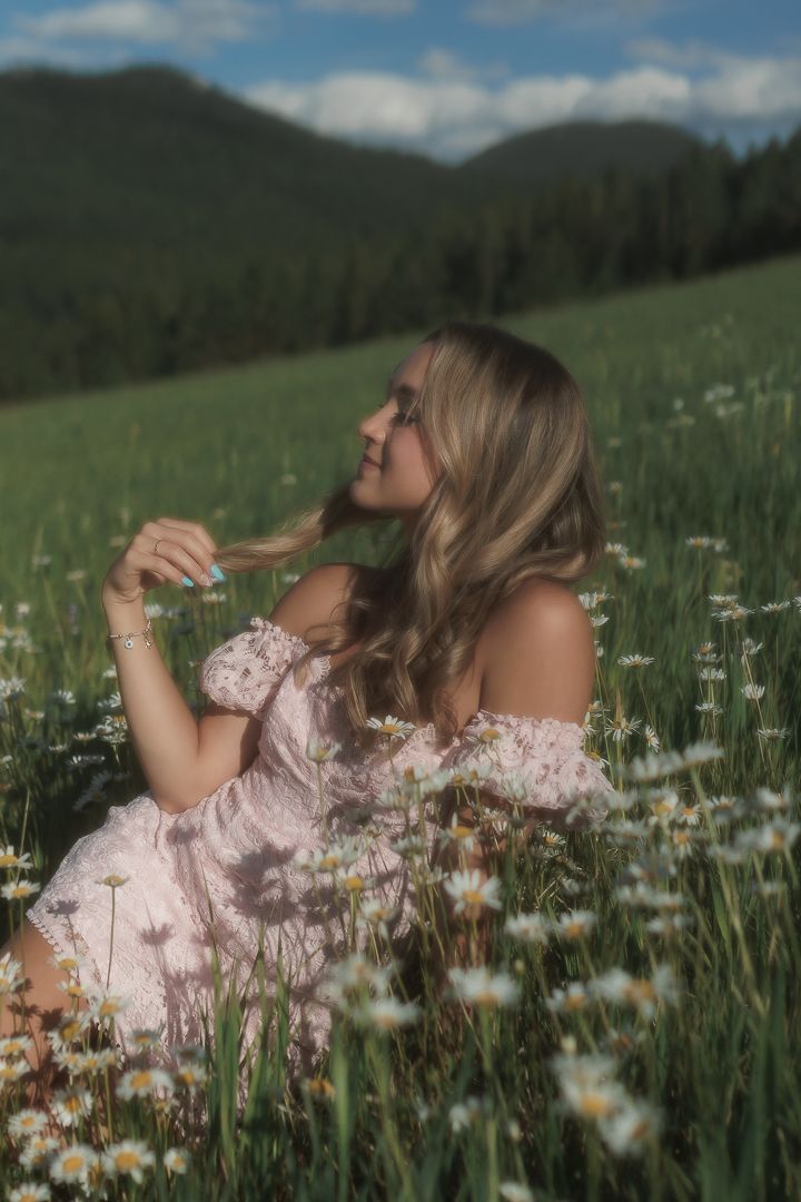 A young woman in a pink off-shoulder dress sits smiling in a field of daisies with mountains behind her.