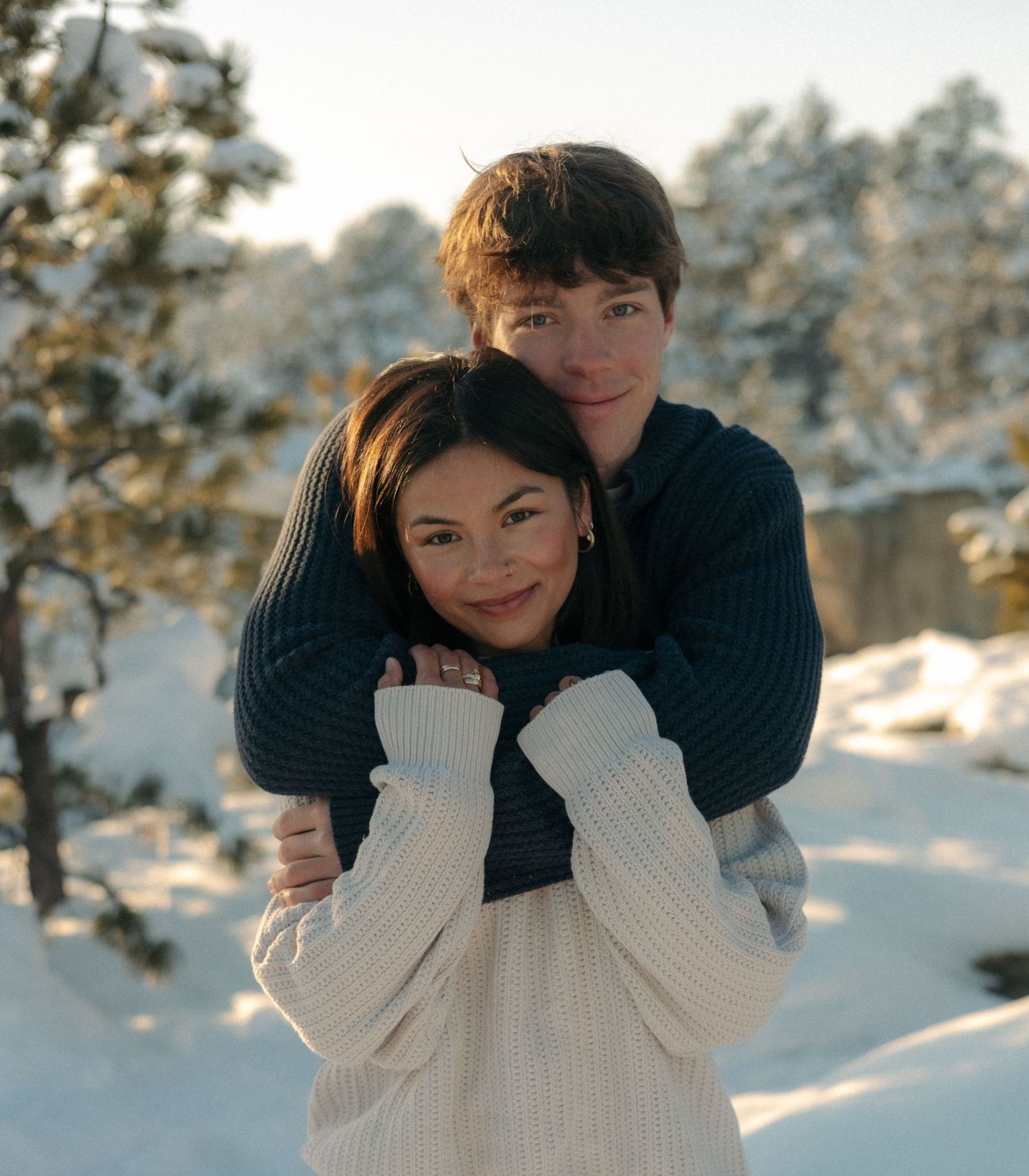 Couple posing together in snowy winter landscape with snow-covered pine trees behind them.