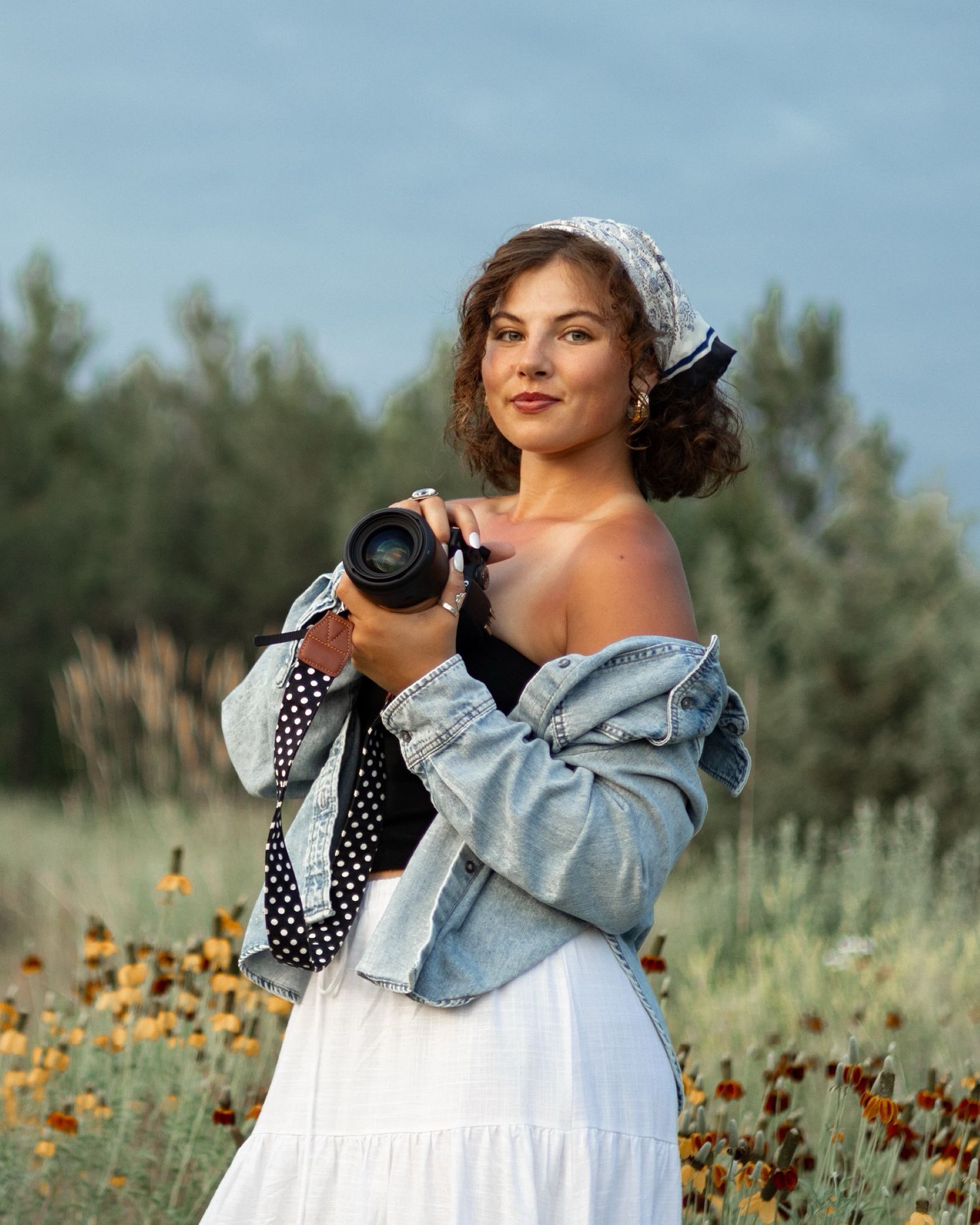 Woman in white skirt and denim jacket holding a camera outdoors in a wildflower field at golden hour.