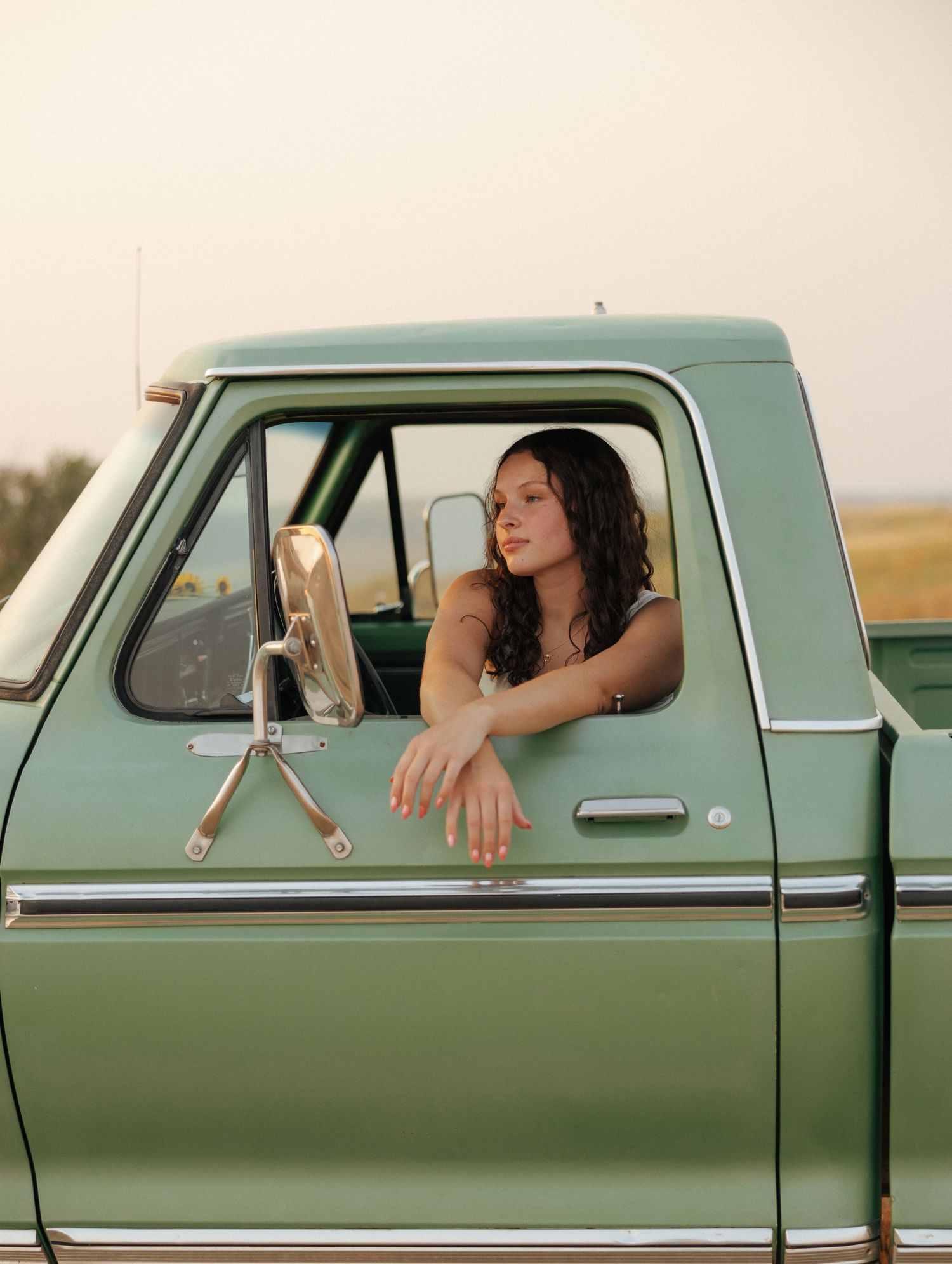 Young woman with curly hair leaning out of a vintage mint green pickup truck window in a rural setting.