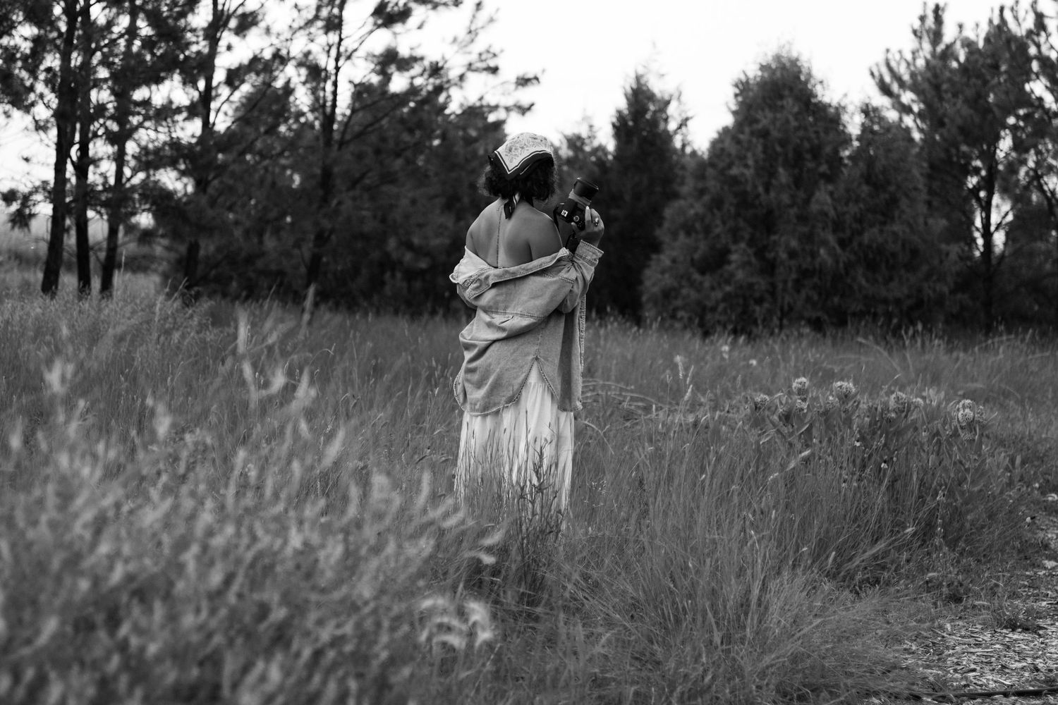 Black and white photo of a woman walking through a grassy field carrying flowers near a forest treeline.
