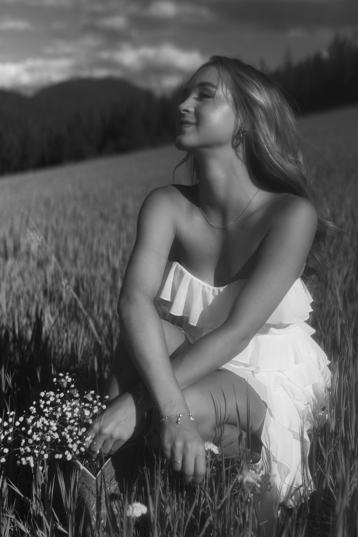 Black and white portrait of smiling young woman kneeling in field holding wildflowers.