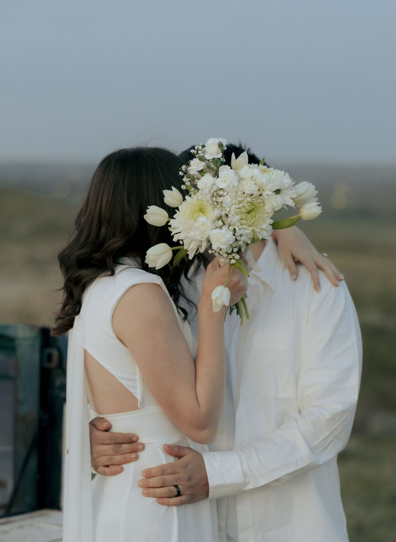 Couple in white attire kissing behind a white floral bouquet of tulips and dahlias outdoors.