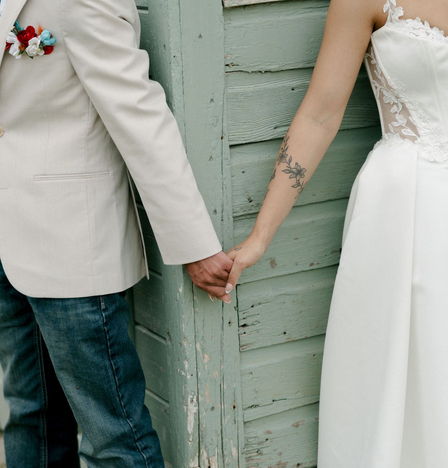 Bride and groom holding hands in front of a rustic green wooden wall, bride wearing lace dress with floral tattoo.