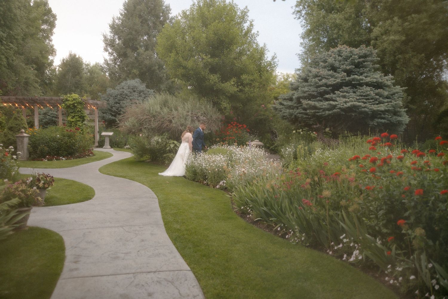 Bride and groom share a romantic moment on a winding garden path surrounded by colorful blooming flowers and lush greenery.