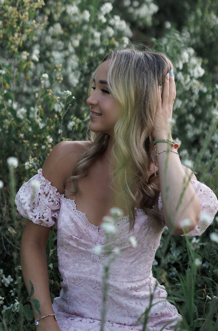 Young blonde woman in pink off-shoulder lace dress smiling among white wildflowers outdoors.