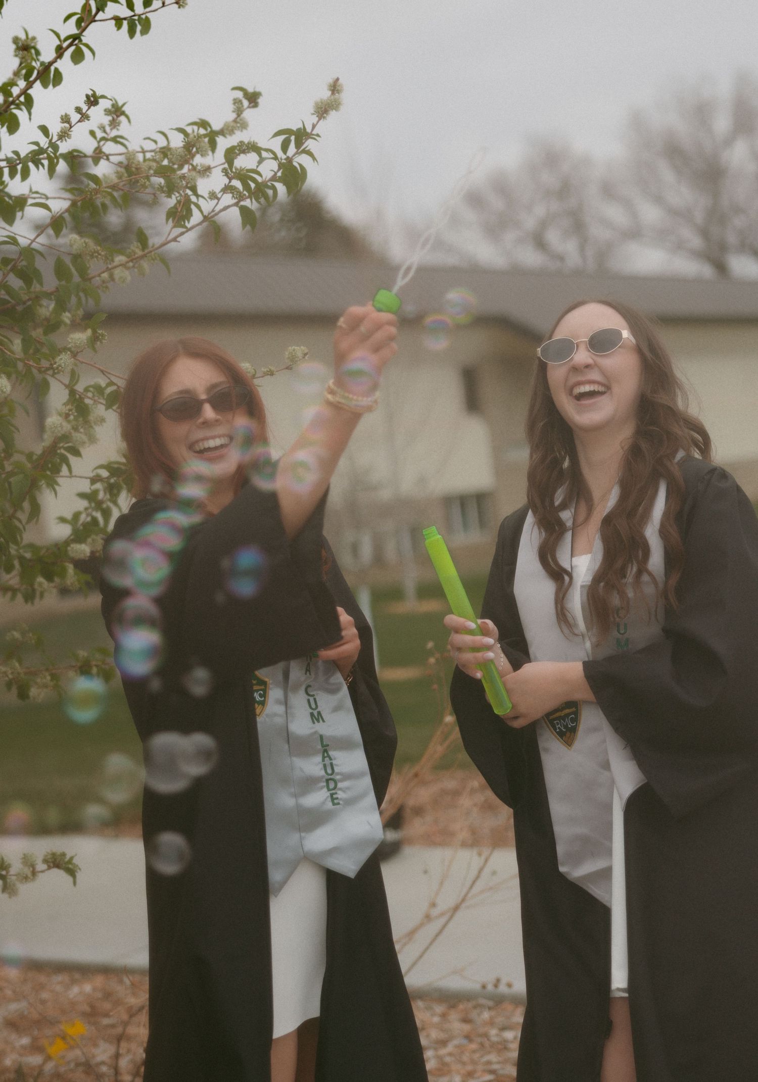 Two graduates in black robes and sunglasses laughing while blowing bubbles outdoors.