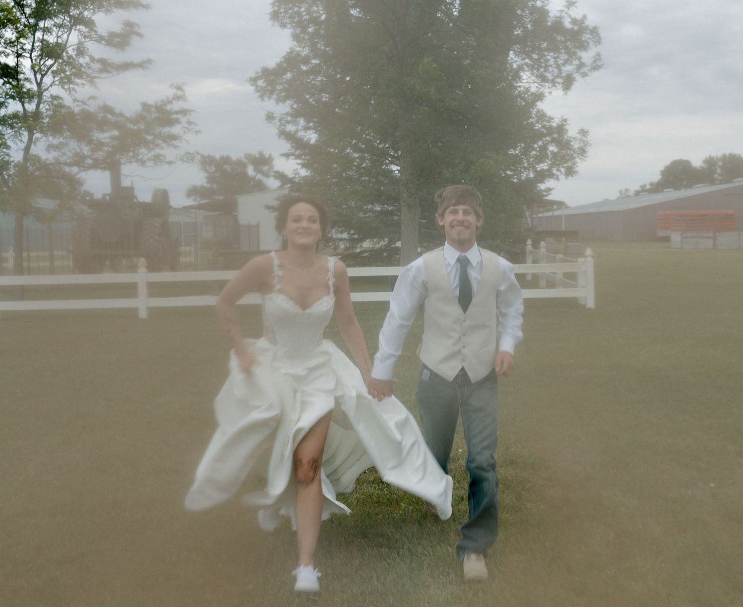Bride and groom running joyfully across green lawn at outdoor wedding venue with white fence and trees.