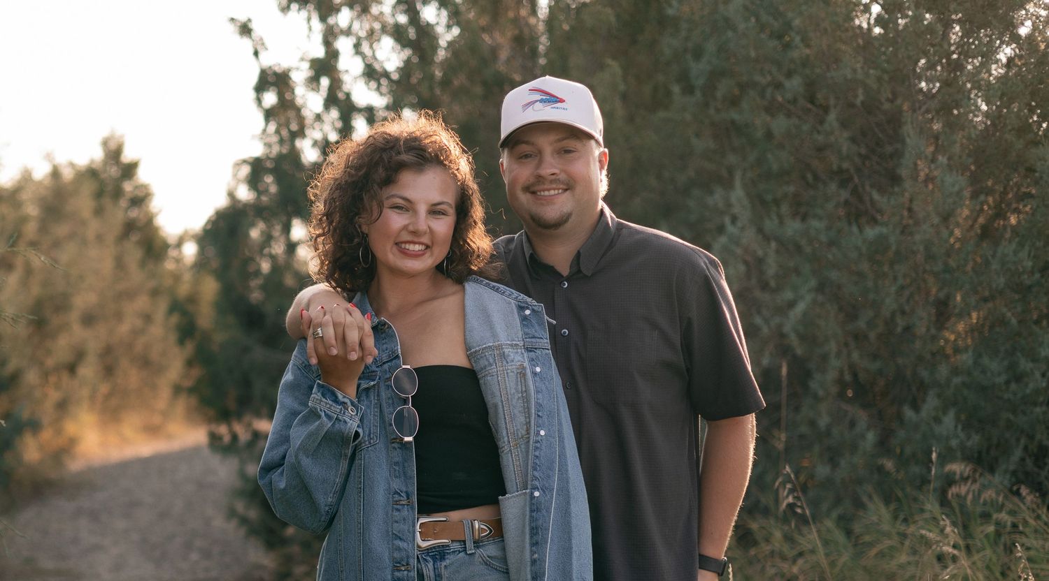 Smiling couple standing together outdoors on a tree-lined path during golden hour engagement photo session.