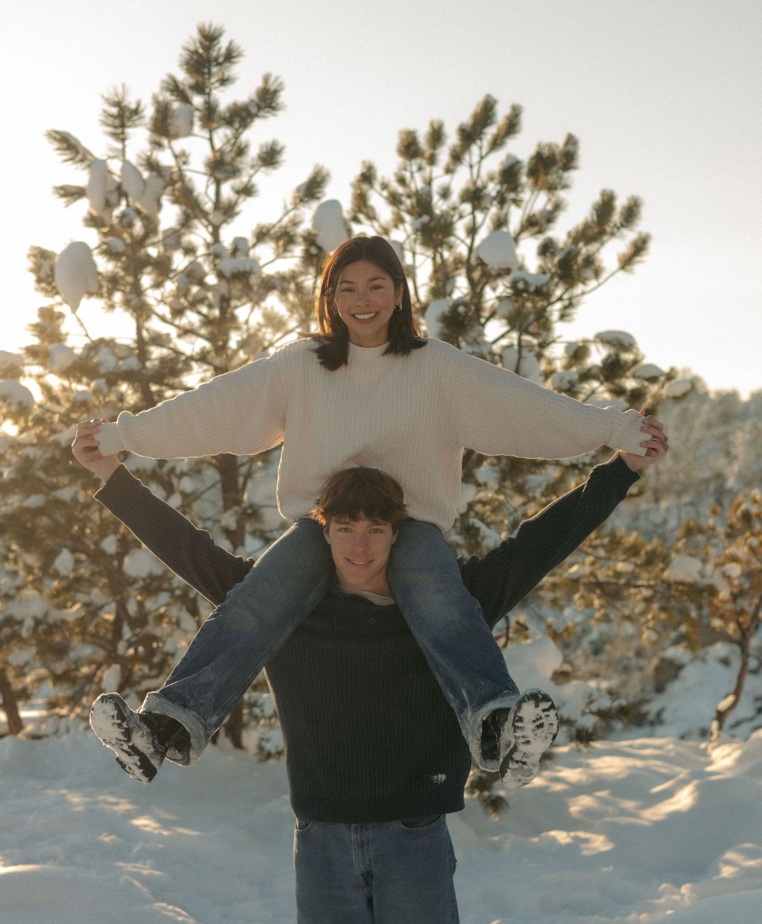 Woman sitting on mans shoulders with arms spread wide in snowy pine forest.