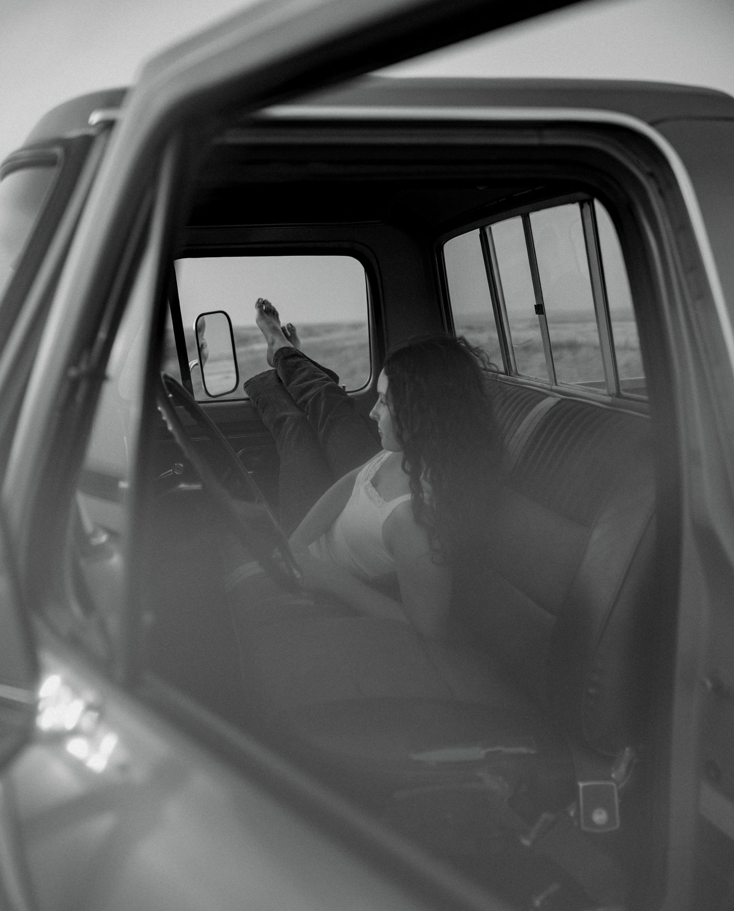 Black and white photo of a woman relaxing in a vintage truck cab with feet up on the dashboard.