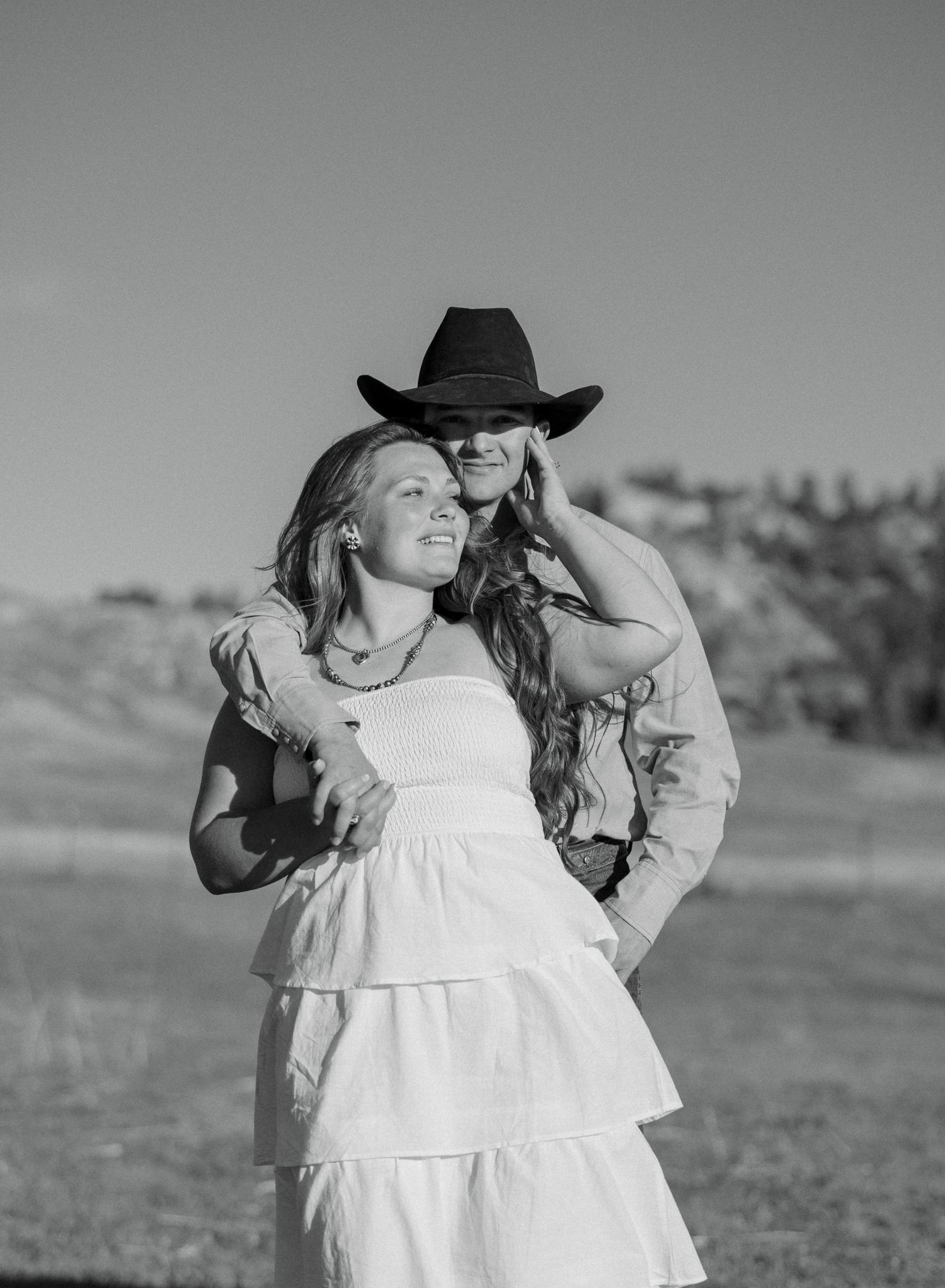 Black and white photo of a couple embracing outdoors, woman in white dress, man in cowboy hat, rural landscape background.