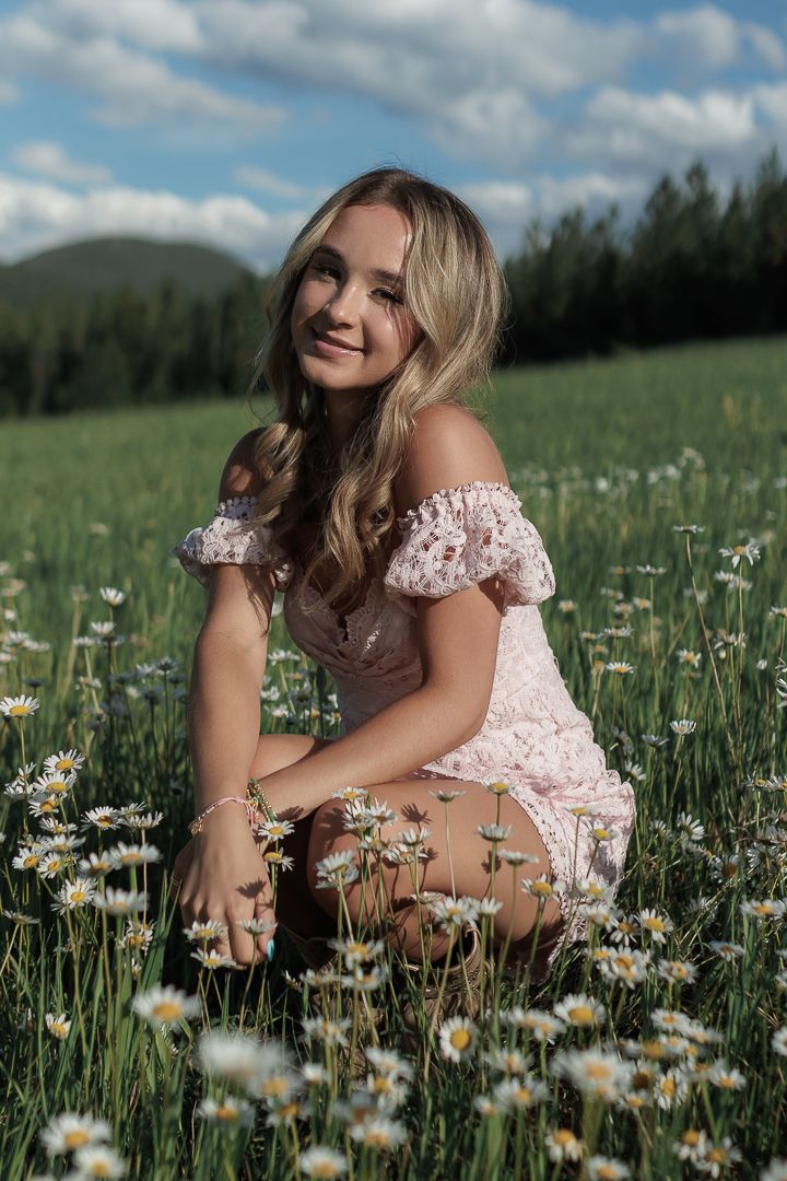 Young blonde woman in pink floral dress crouching in daisy field under blue sky.
