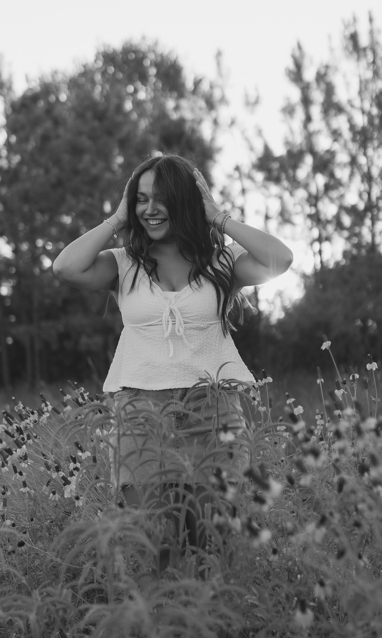 Black and white portrait of smiling woman standing in wildflower meadow with trees behind.