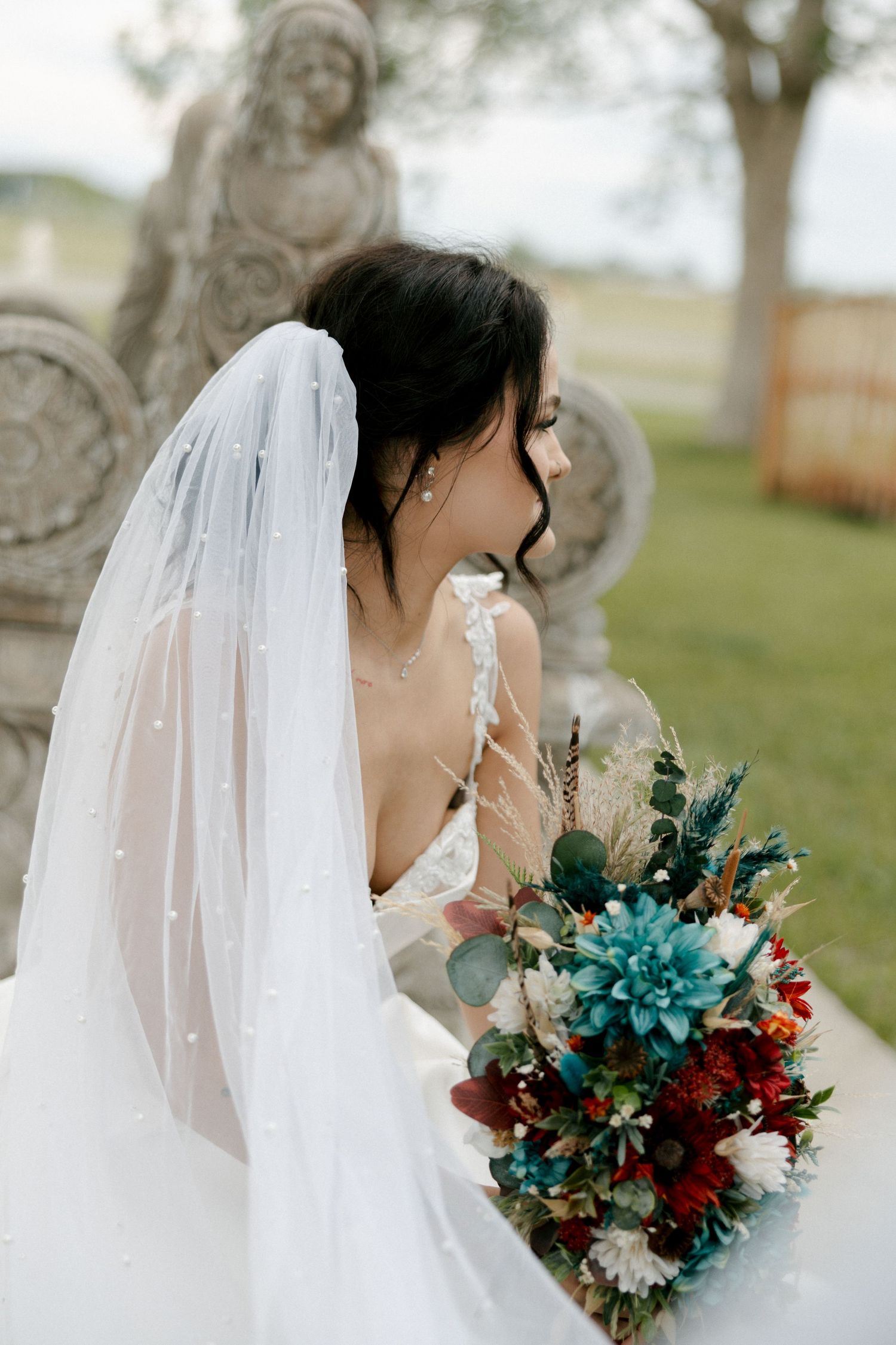 Bride in white gown and pearl veil holds colorful bouquet with teal, red, and white flowers beside stone sculptures.