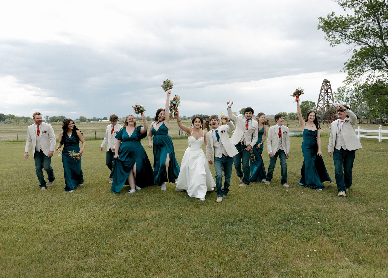 Wedding party in teal dresses and white suits celebrating outdoors on green lawn with raised arms and bouquets.
