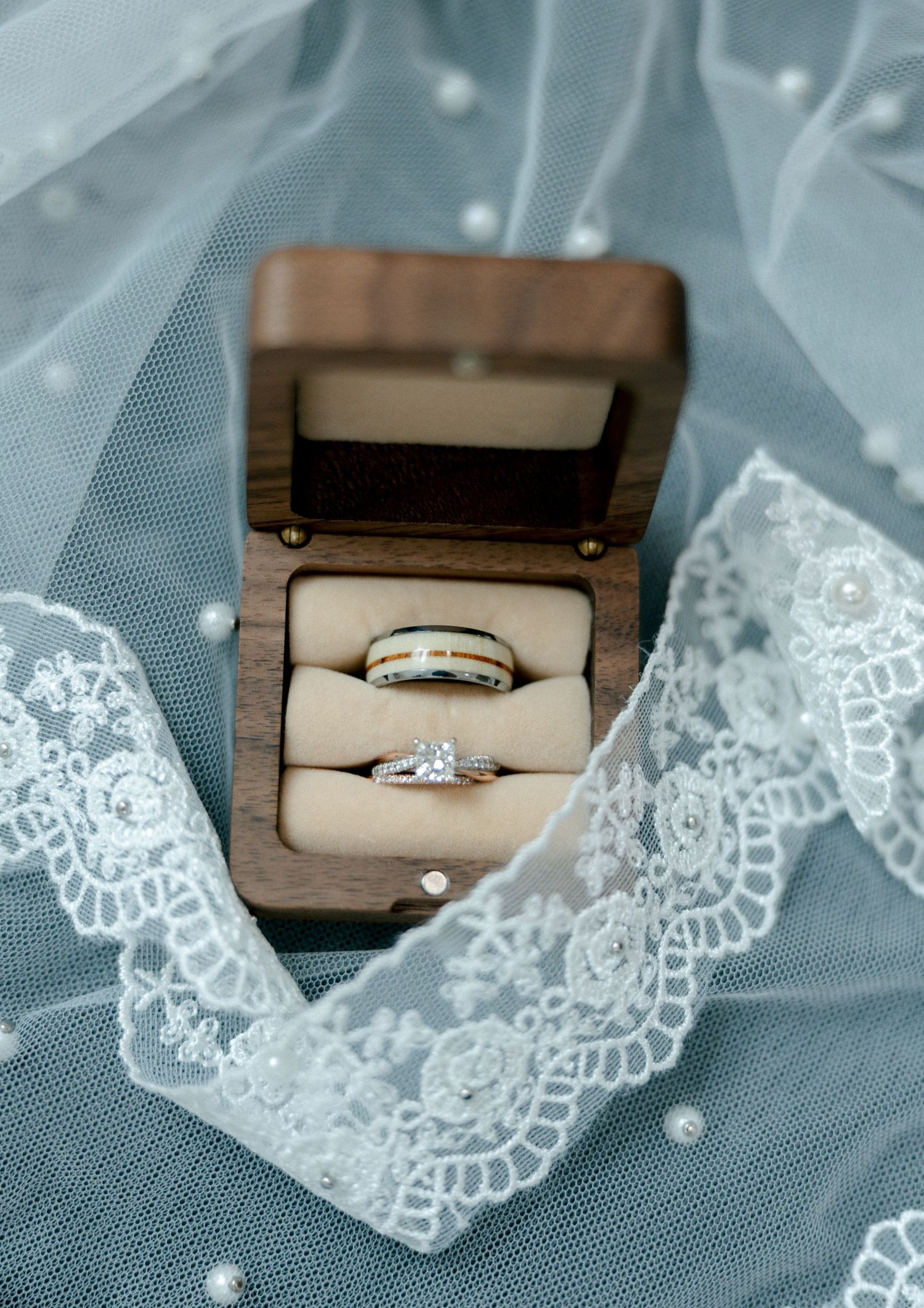 Wedding rings in a wooden box on a lace bridal veil, featuring a diamond engagement ring and a gold band.