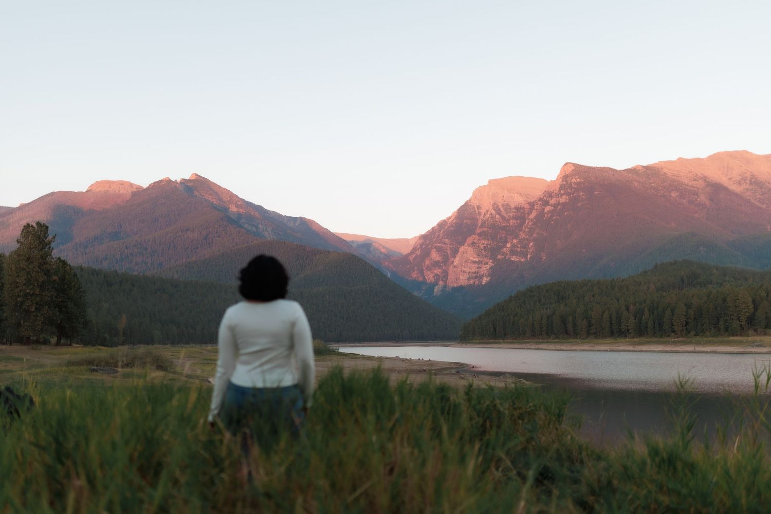 Woman in white top stands with her back to camera gazing at pink alpenglow on rugged mountain peaks above a lake.