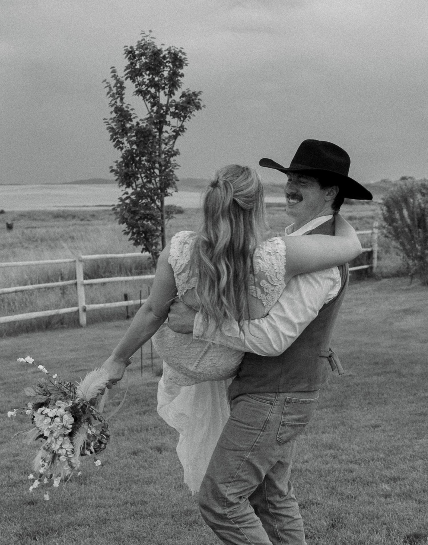 Black and white photo of a couple dancing outdoors at a rustic wedding, bride holding bouquet, groom in cowboy hat.
