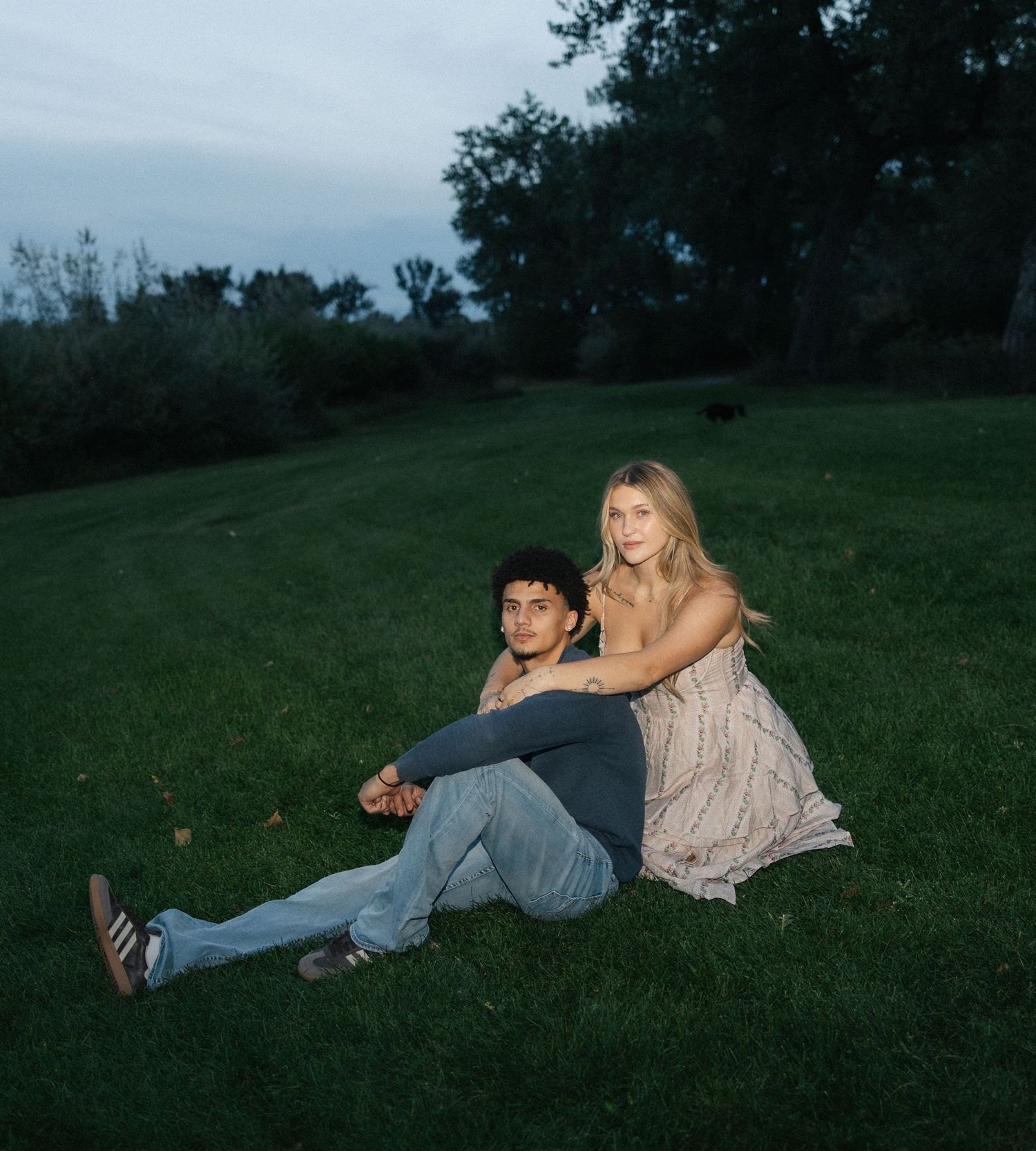 A young couple sits together on a lush green lawn at dusk, surrounded by trees in a park setting.