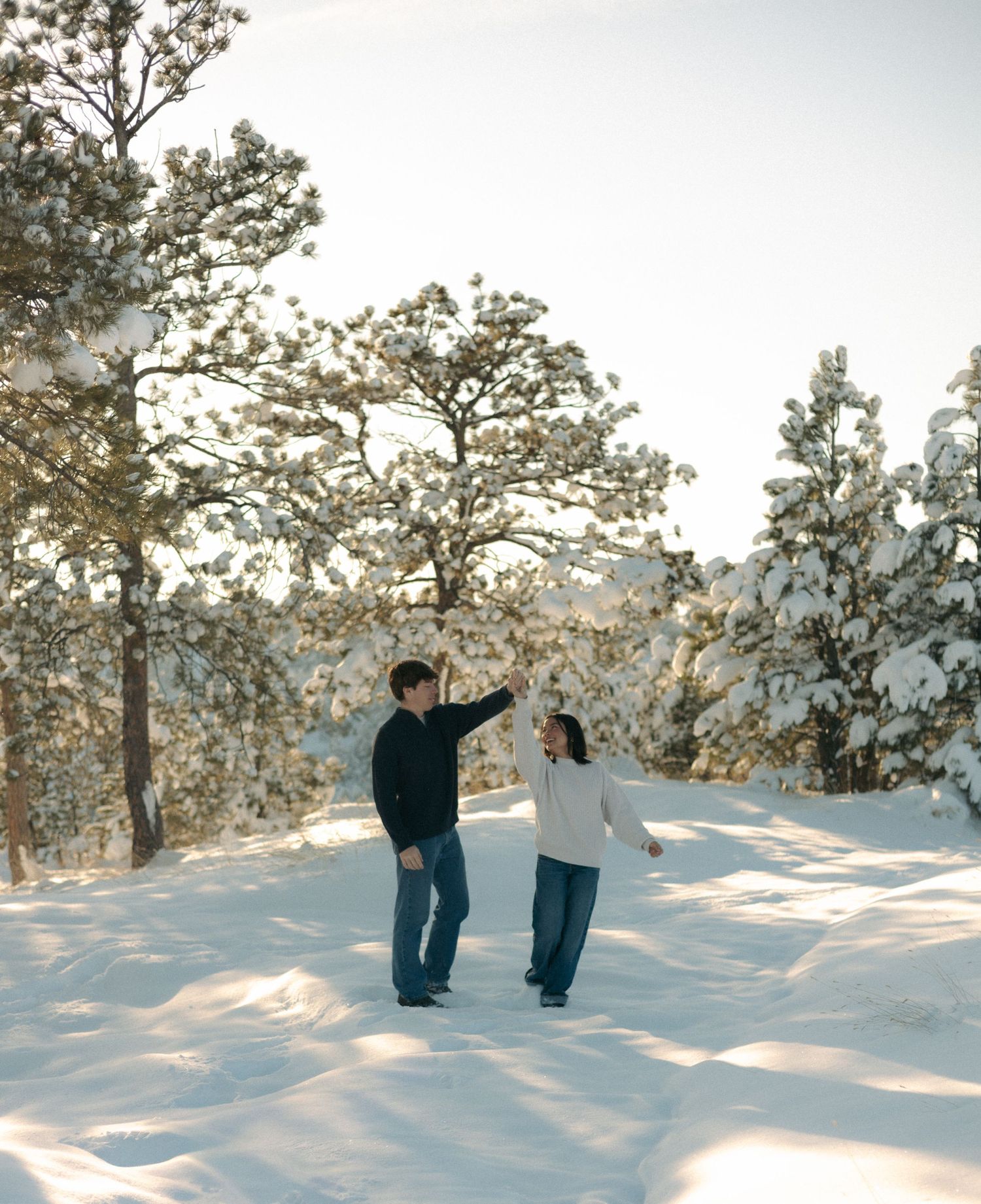 Couple dancing playfully in snowy winter forest with snow-covered pine trees in sunlight.
