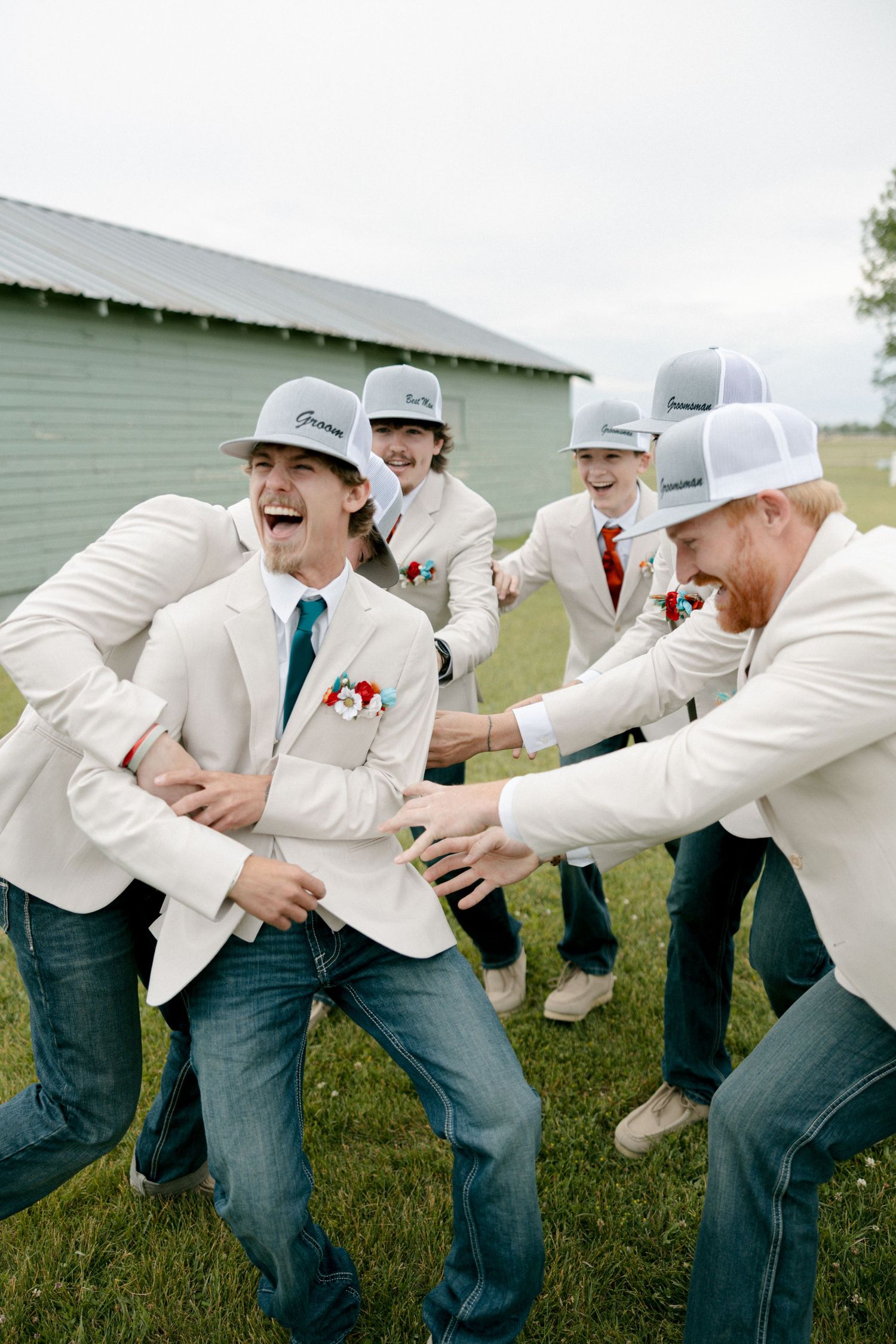 Groomsmen in white blazers, jeans, and trucker hats laughing and joking around outside a green barn.