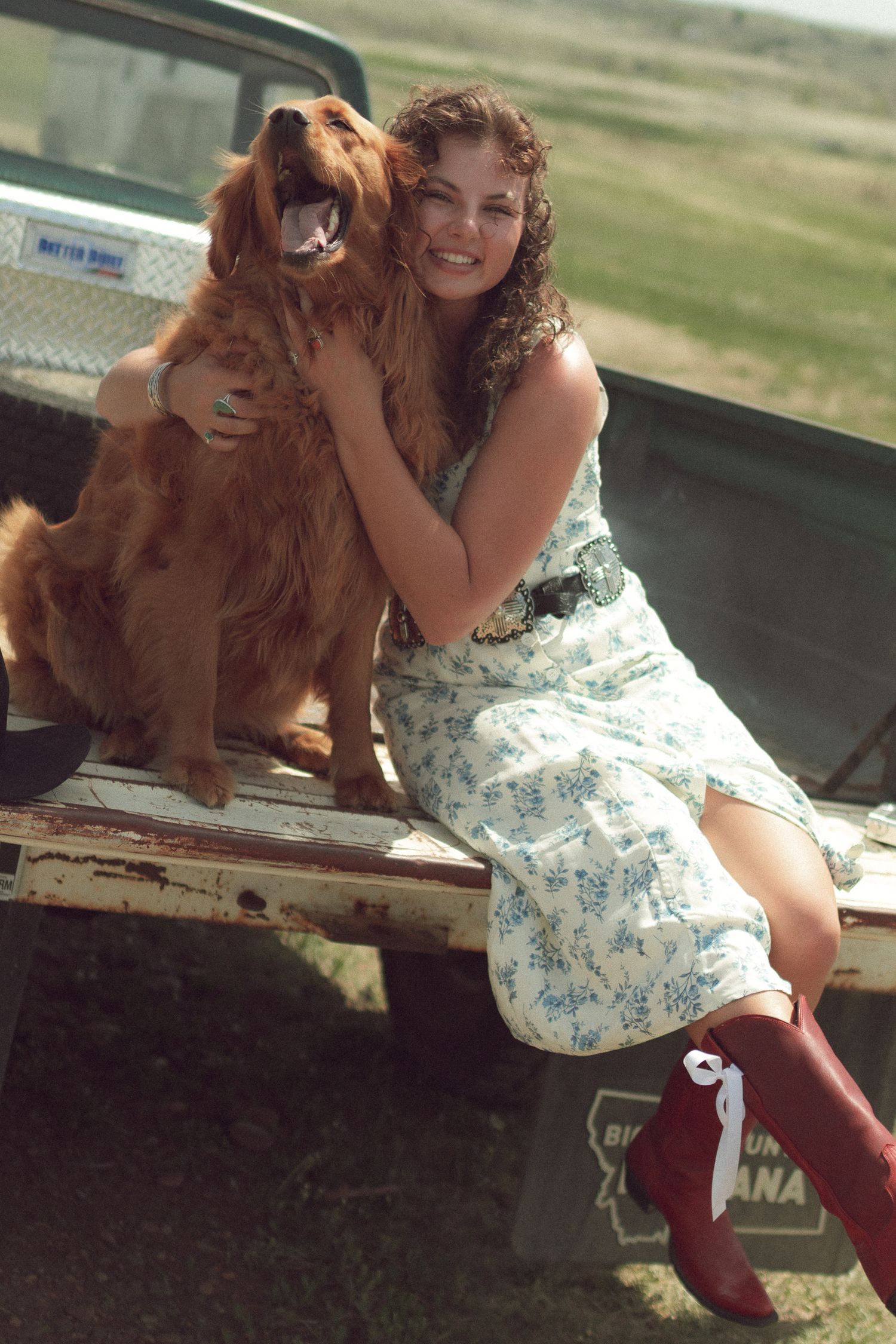 Smiling woman in floral dress and red boots sitting on truck tailgate hugging a happy golden retriever dog.