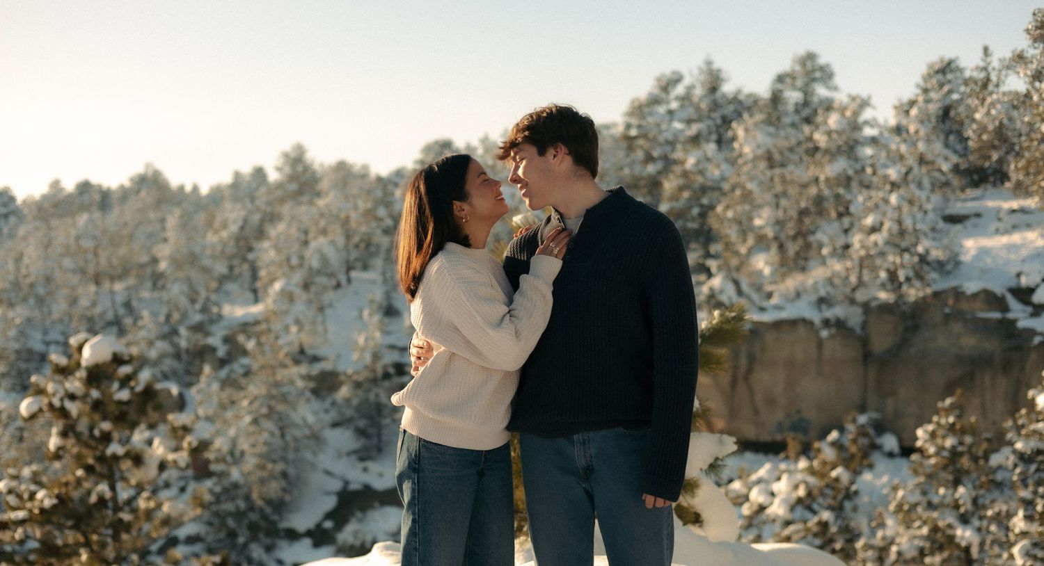 Couple leaning in for a kiss surrounded by snow-covered trees in winter sunlight.