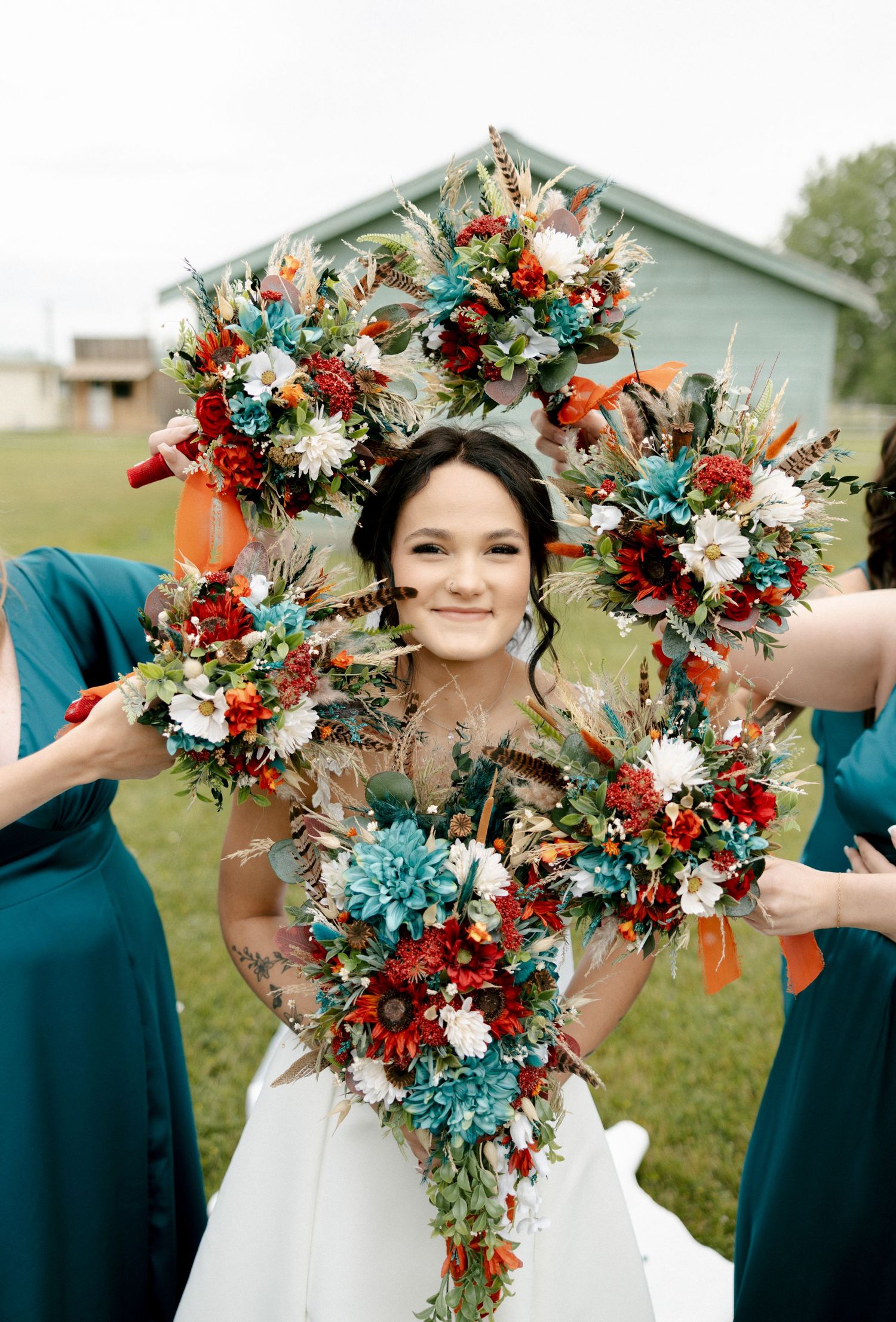 Smiling bride framed by a large floral wreath with teal, red, and white blooms held by bridesmaids in teal dresses.