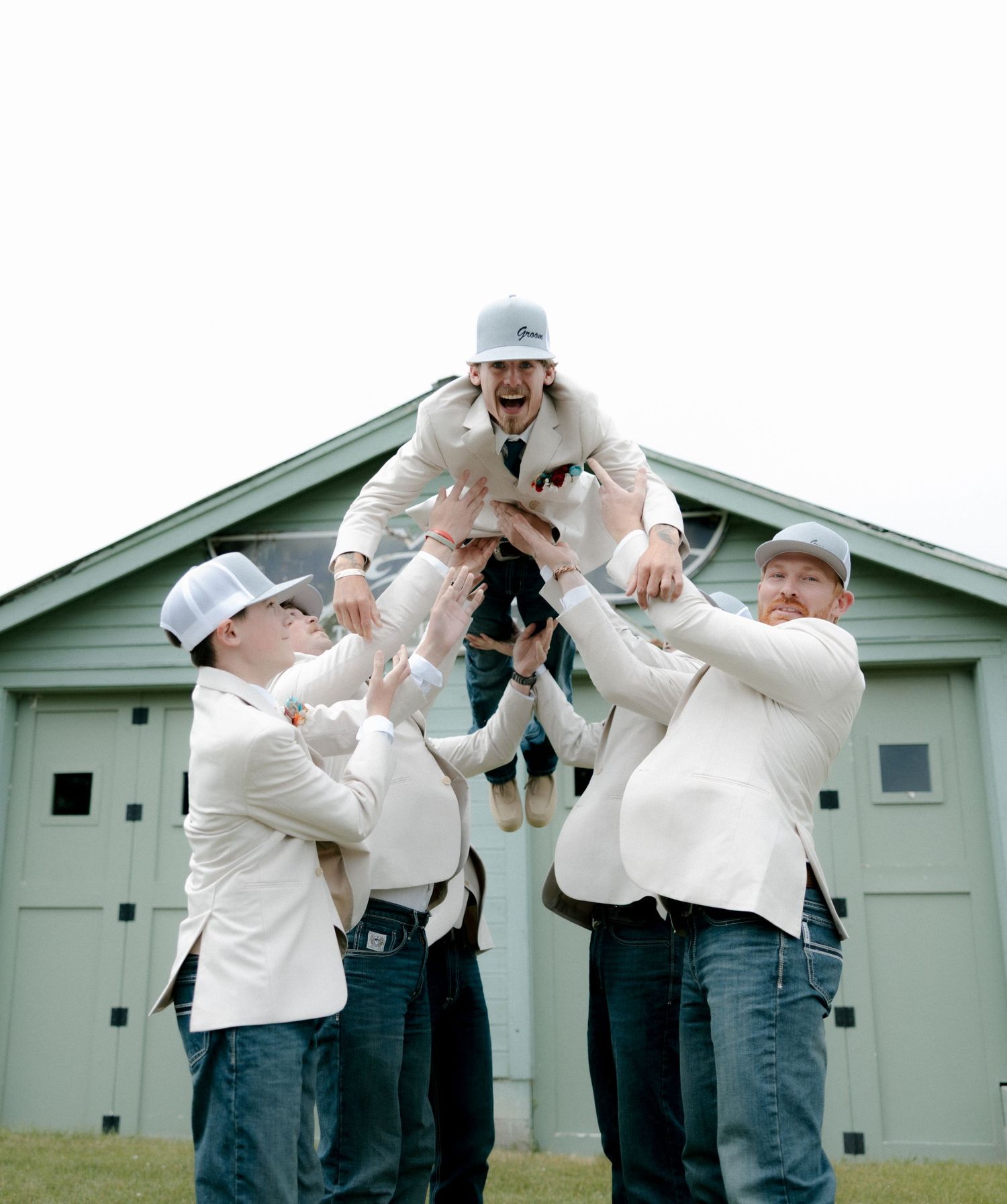 Groomsmen in white jackets and caps tossing groom in air outside barn at wedding celebration.