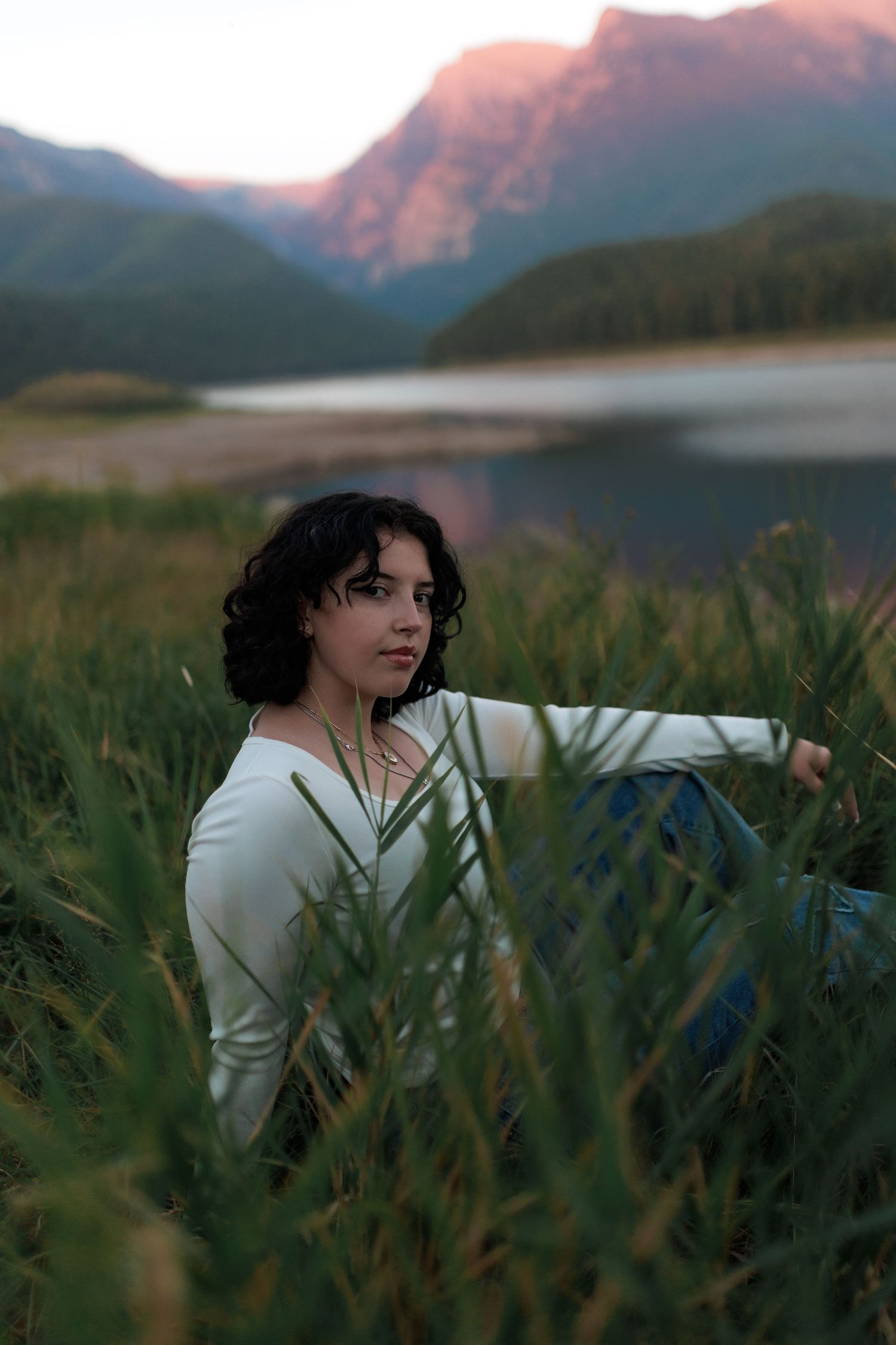 Young woman with curly dark hair sits in tall grass by a mountain lake at sunset wearing a white long-sleeve top.
