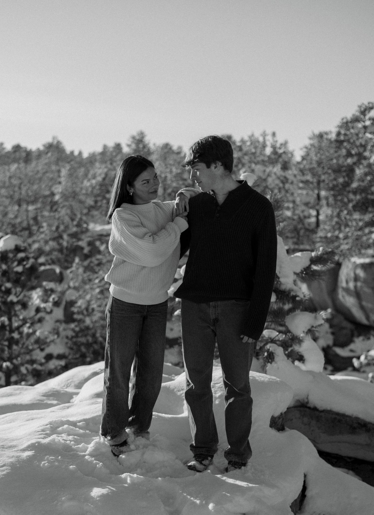 Black and white photo of couple holding hands on snowy rocks with pine trees.