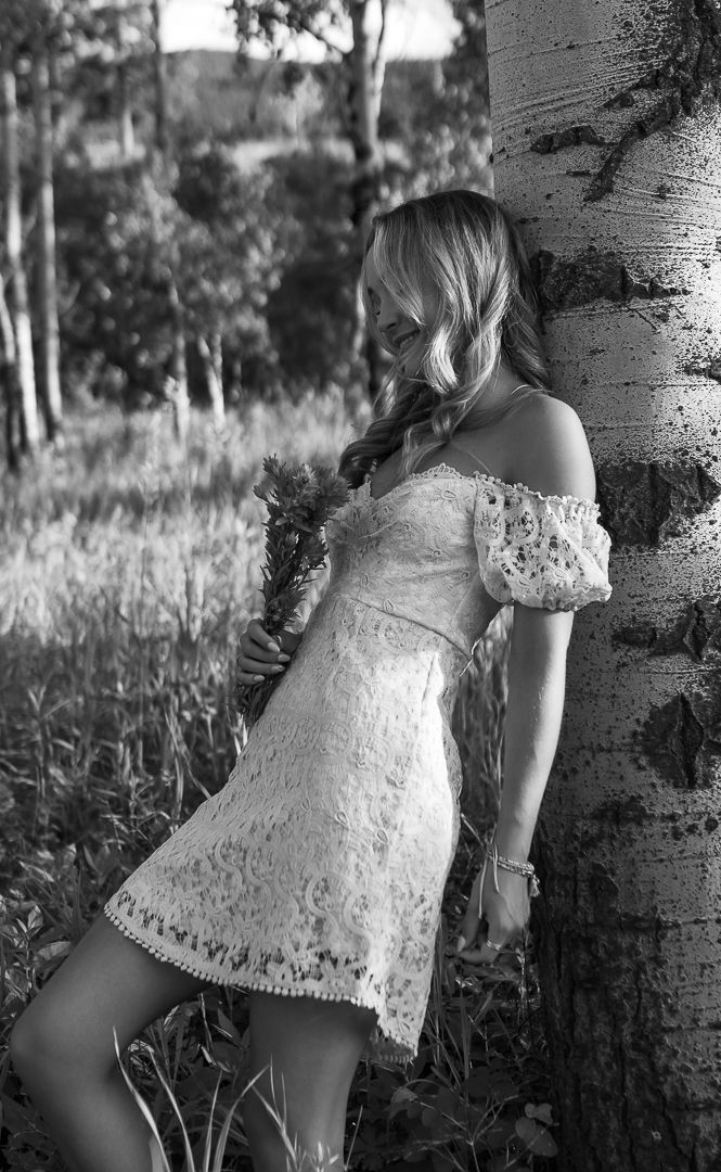 Black and white photo of young woman in lace off-shoulder dress leaning against birch tree.