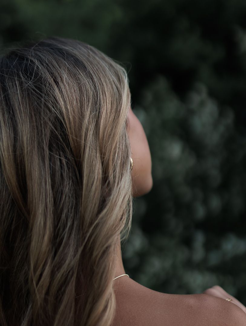 Close-up profile of woman with long highlighted blonde hair against dark green foliage background.