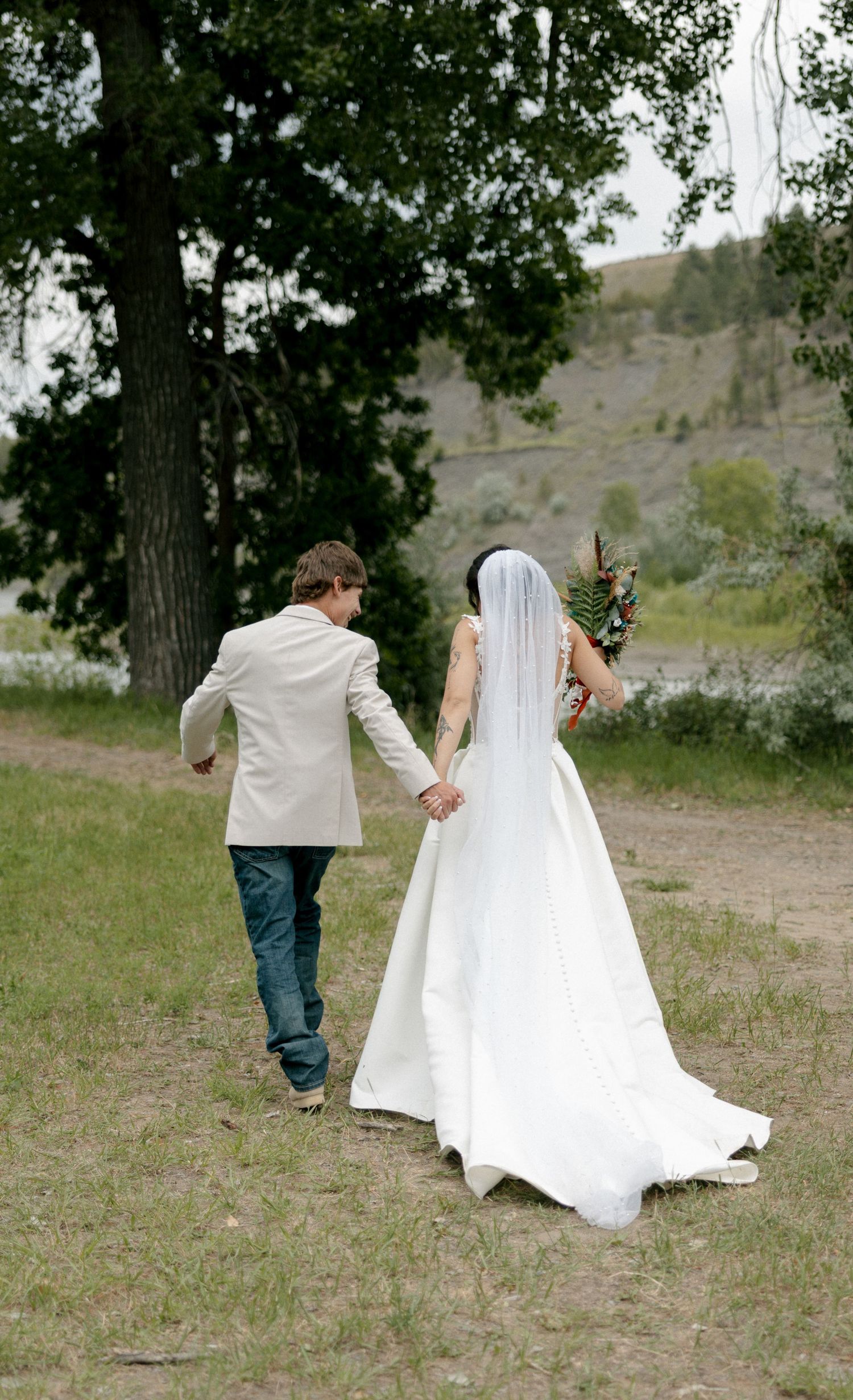 A bride and groom walk hand-in-hand through a scenic outdoor setting, with the bride in a white gown and veil.