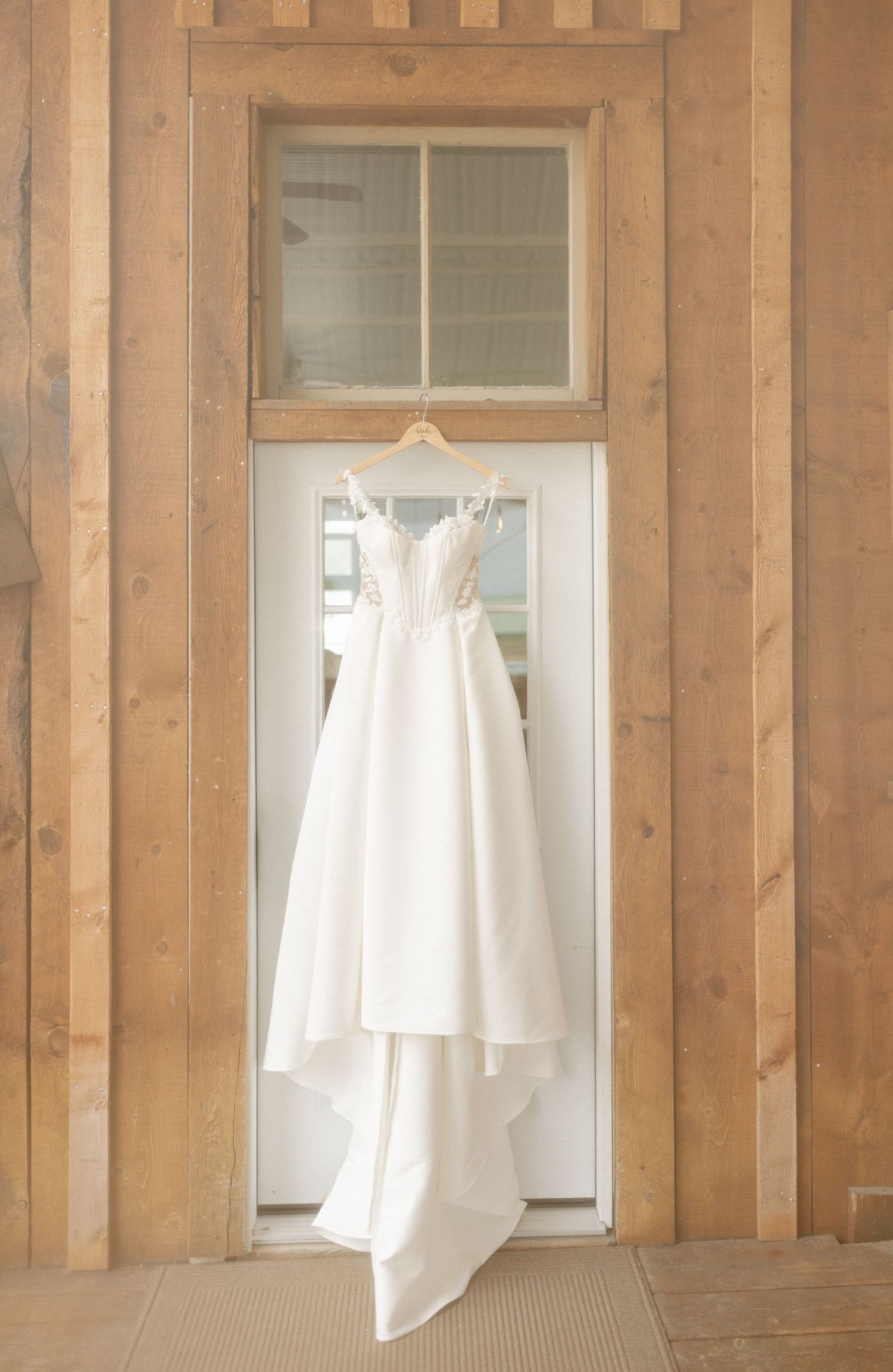 White wedding dress hanging on a wooden door frame in a rustic barn setting with natural light.