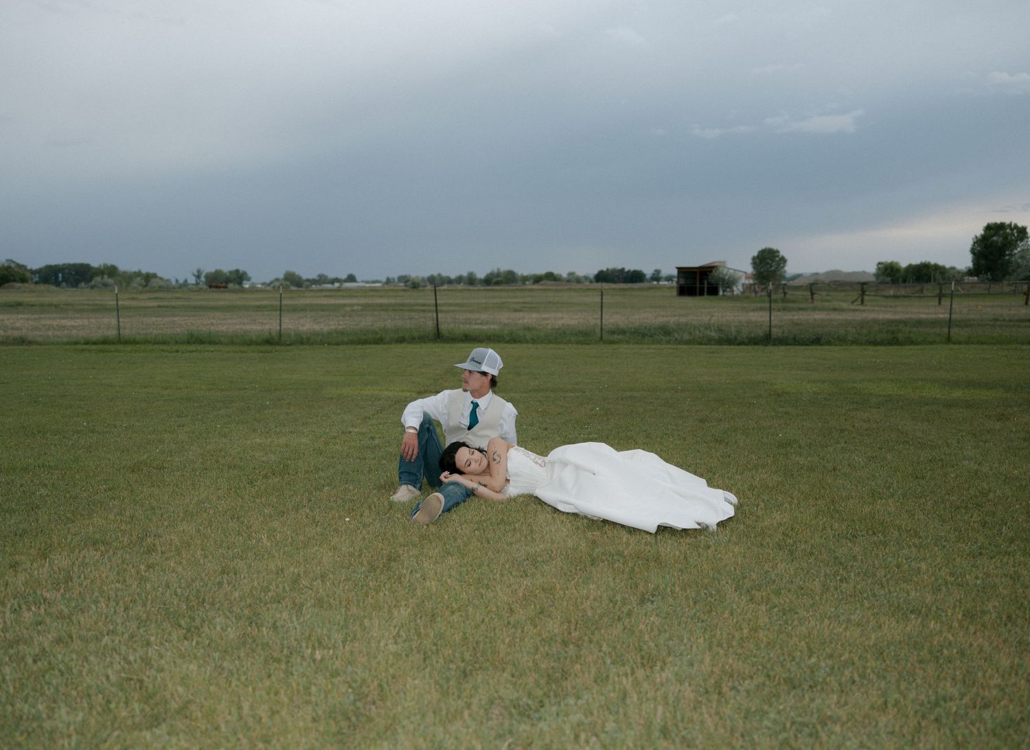 Bride in white gown lying in grooms lap on green field under dramatic cloudy sky.
