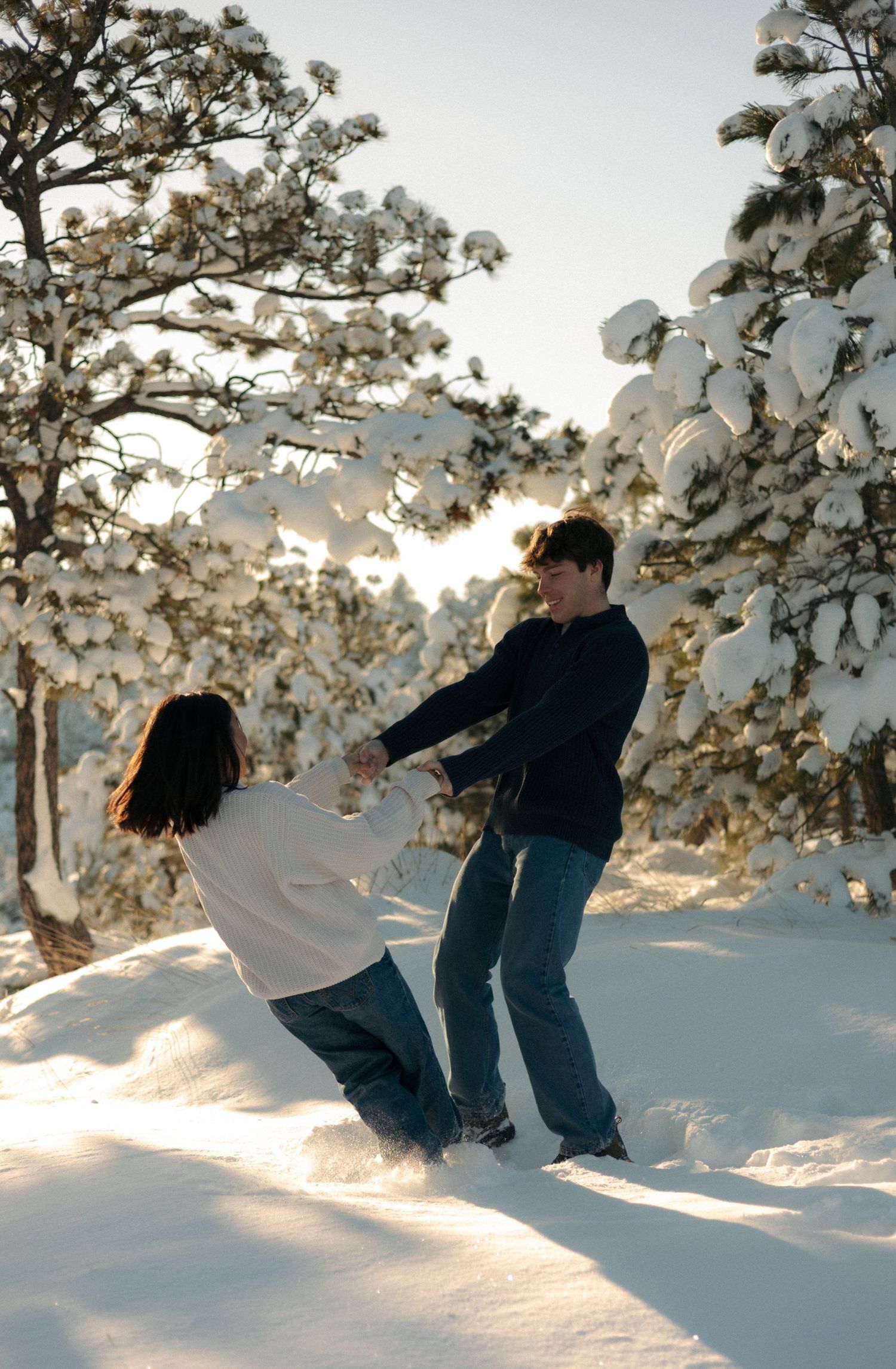 A couple playfully holds hands and dances together in deep snow surrounded by winter pine trees.