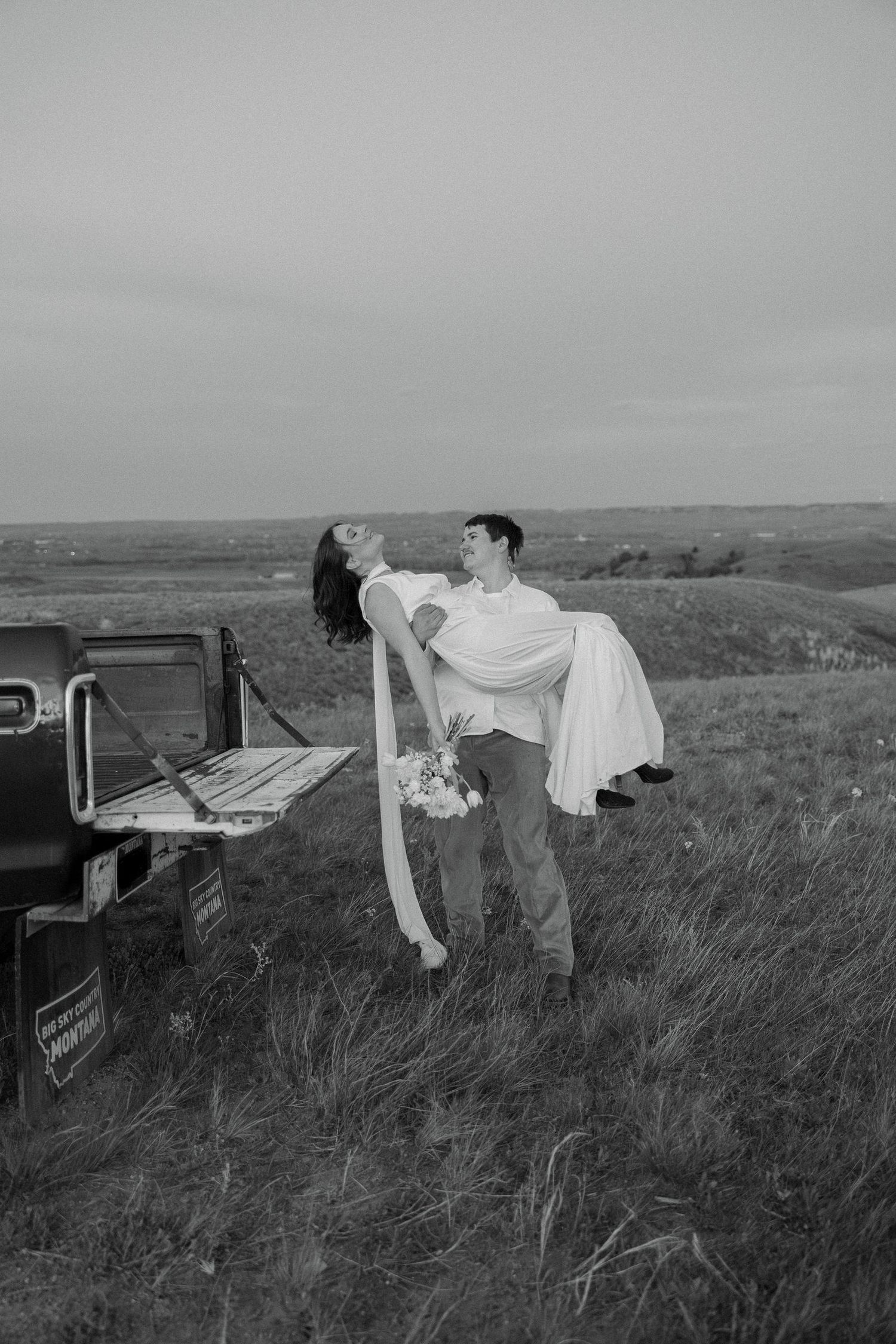 Black and white photo of groom carrying bride in field near vintage truck during romantic outdoor wedding shoot.