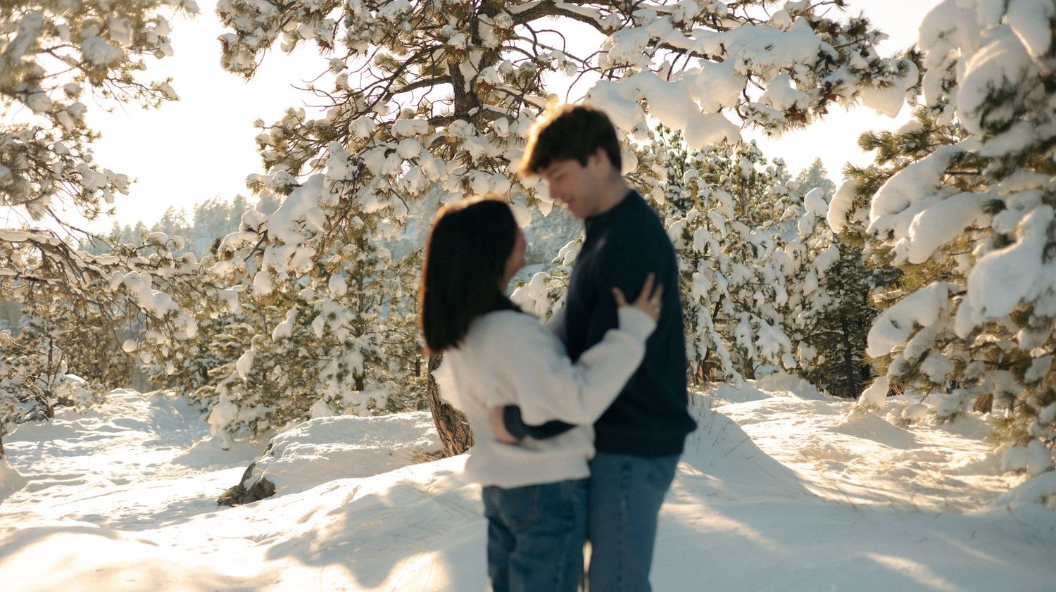Romantic couple embracing and nearly kissing in a sun-drenched snowy forest with frosted pine trees.