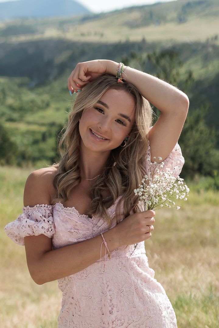 Smiling young woman in pink off-shoulder lace dress holding wildflowers with mountain backdrop.