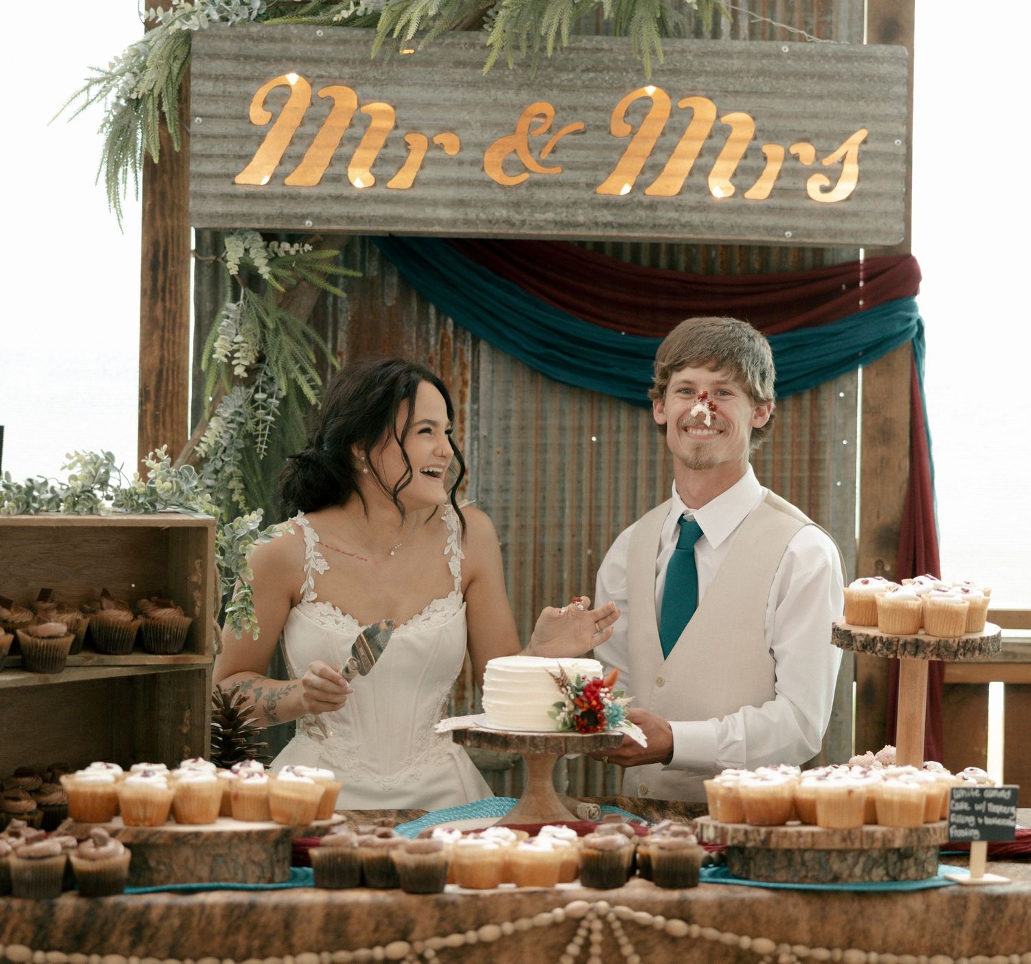 Bride and groom cutting wedding cake at rustic dessert table with cupcakes under Mr & Mrs sign.
